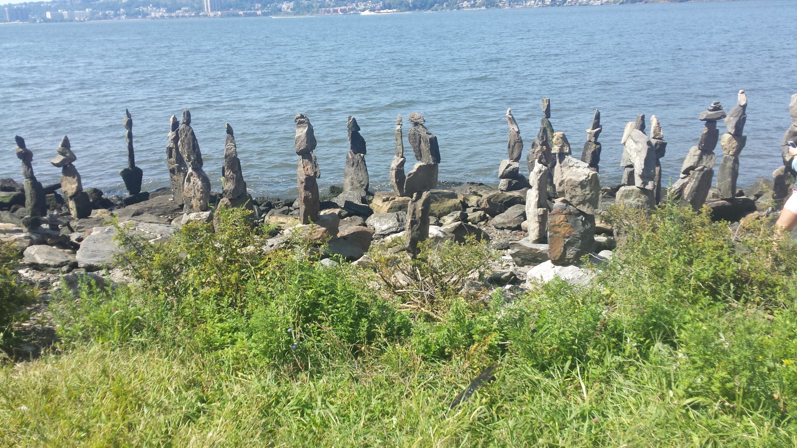 A line of carefully balanced stones arranged vertically along a rocky shoreline, with a body of water in the background and greenery in the foreground. The scene captures a sunny day with clear skies. West Street Greenway mountain bike trail.