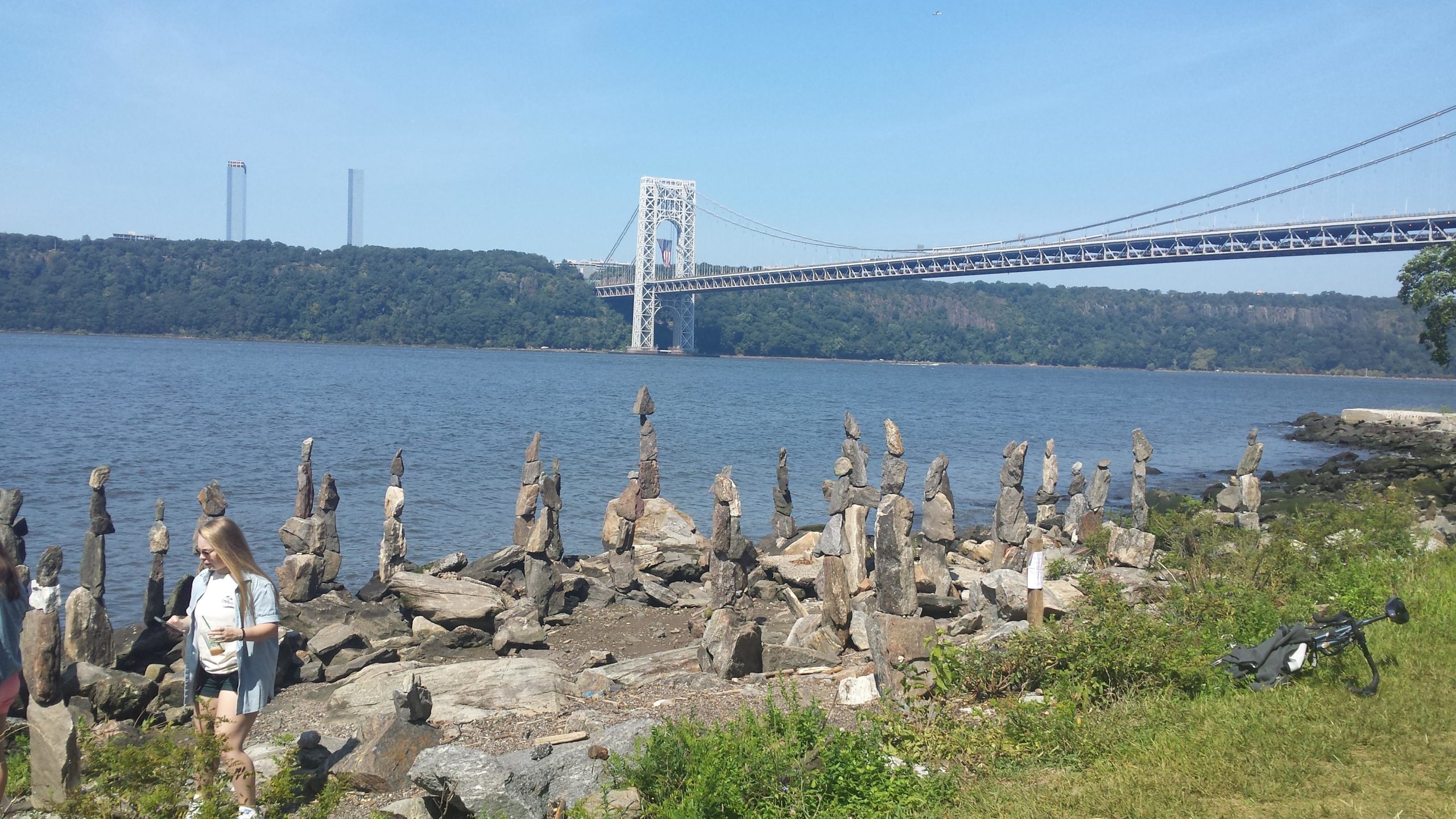 A view of the Hudson River with the George Washington Bridge in the background. In the foreground, several stacked rock formations are arranged along the shoreline, while a person walks nearby. The scene is set against a clear blue sky and green hills. West Street Greenway mountain bike trail.