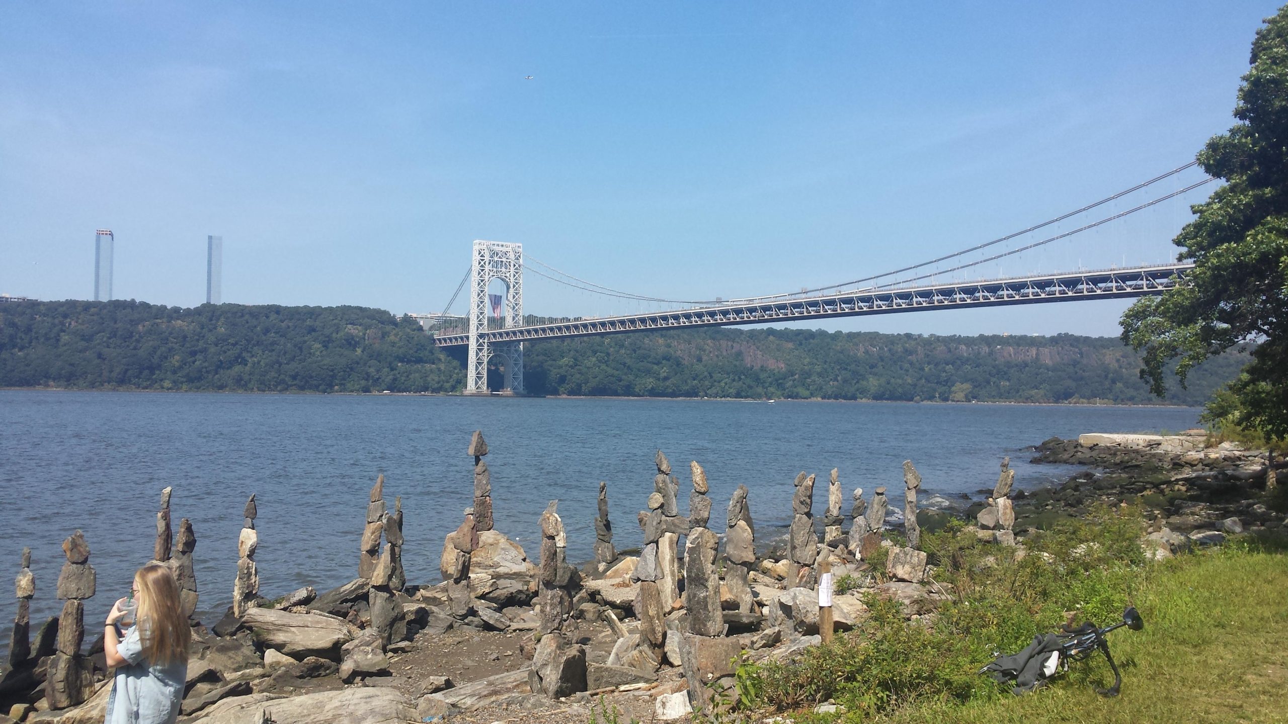 A scenic view of the George Washington Bridge spanning over the Hudson River, with a rocky shoreline in the foreground featuring several stacked stones. A person with long hair is seen facing away from the camera, standing beside the water, while lush greenery and distant hills are visible in the background under a clear blue sky. West Street Greenway mountain bike trail.