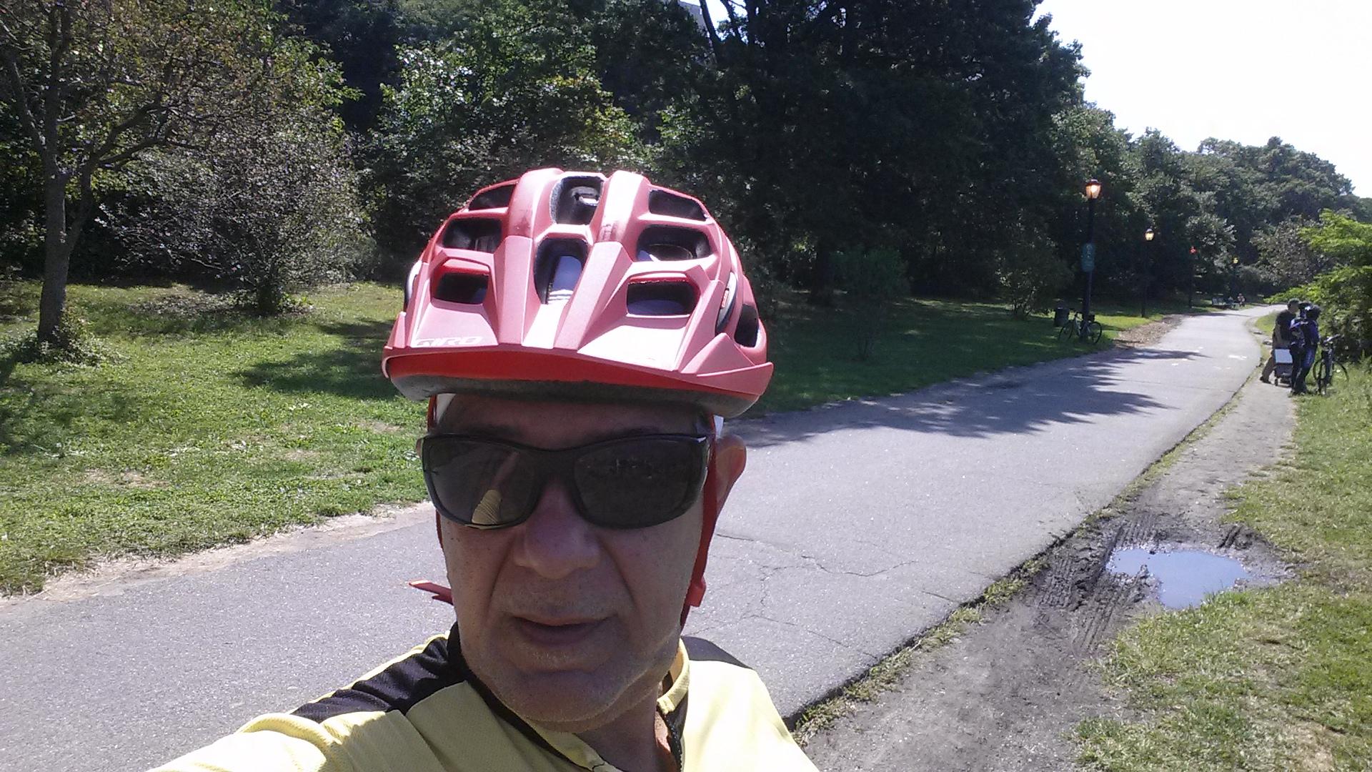 Alt tag: "A person wearing a red cycling helmet and sunglasses takes a selfie while standing on a bike path in a sunny park. Green trees and grass are visible in the background." West Street Greenway mountain bike trail.