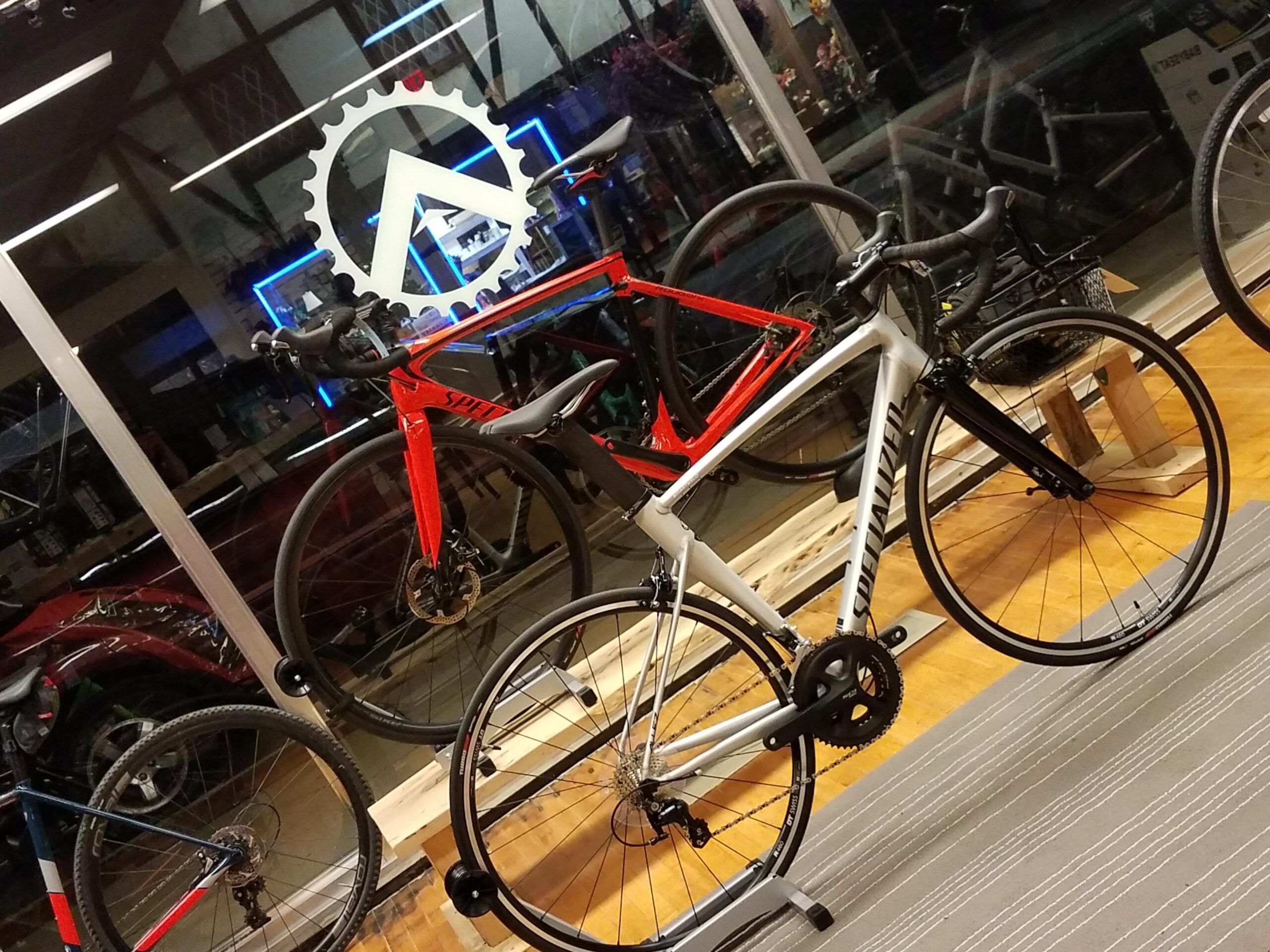 Image of a bike shop interior featuring two bicycles on display: one is a silver road bike with black accents, and the other is a red racing bike. A large logo featuring a gear design is visible in the background, illuminated by blue lighting. The shop's wooden flooring and striped rug add to the inviting atmosphere.
