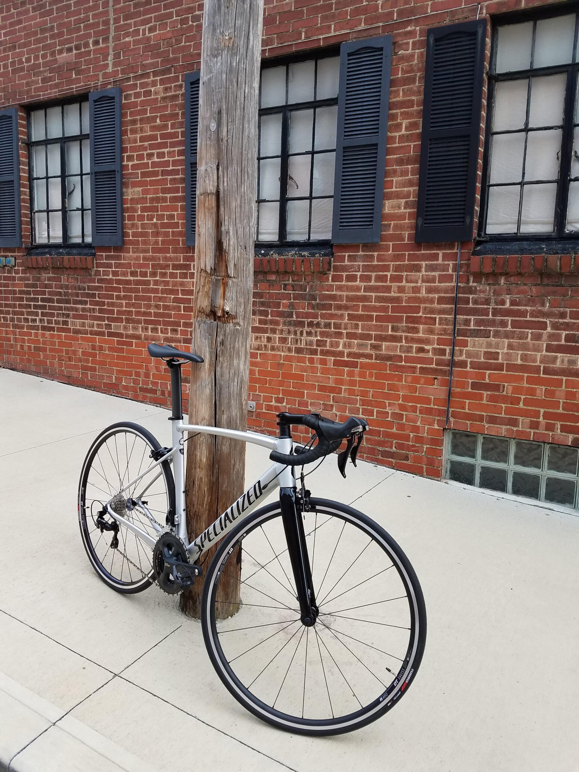 A silver and black road bike leaning against a wooden utility pole, positioned on a concrete sidewalk next to a brick wall with black-shuttered windows.