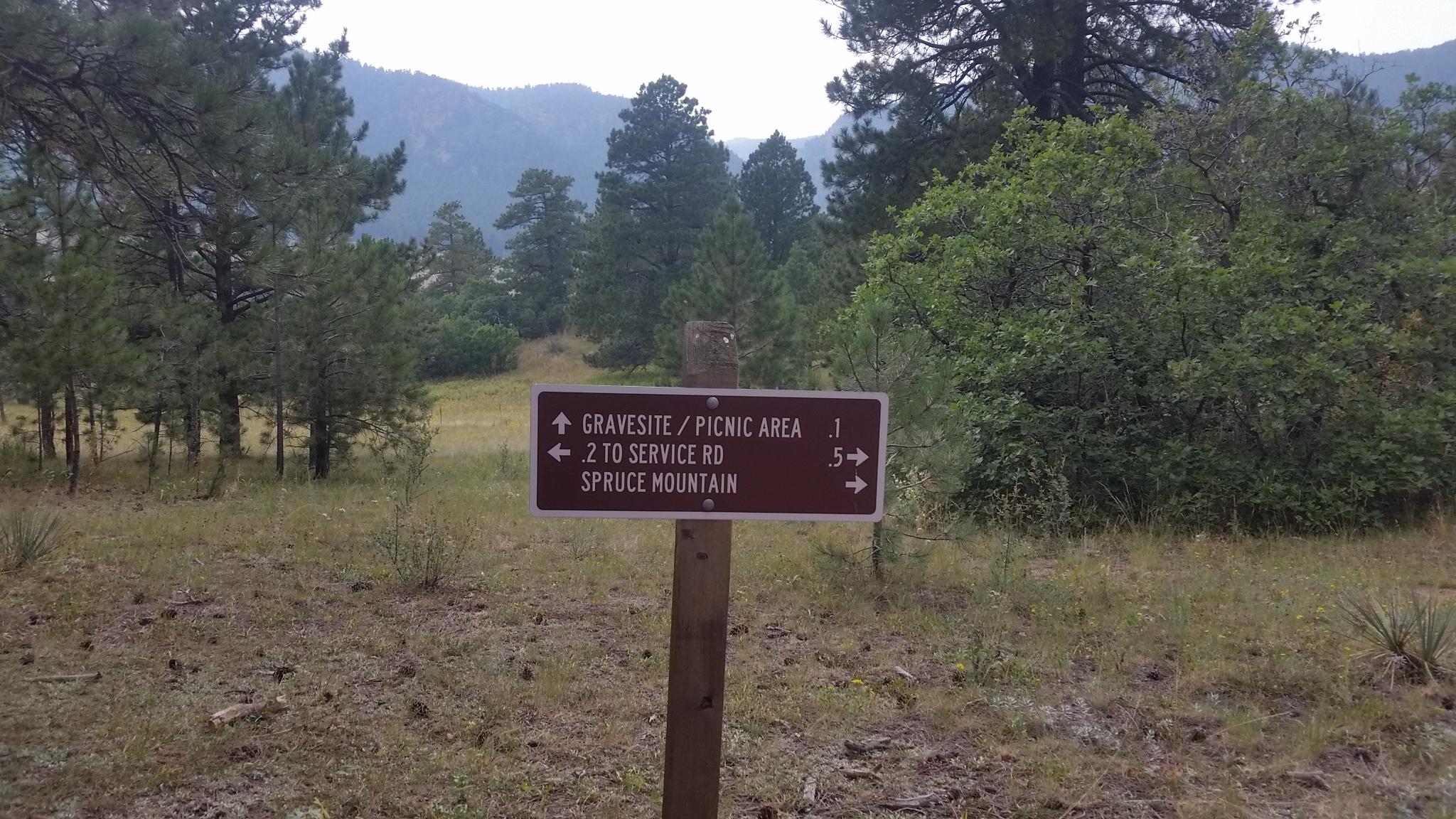 A wooden signpost in a natural setting, indicating directions to a gravesite/picnic area, service road, and Spruce Mountain, with distances marked in miles. Surrounding greenery includes tall trees and shrubs. Spruce Mountain Trail Upper Loop mountain bike trail.