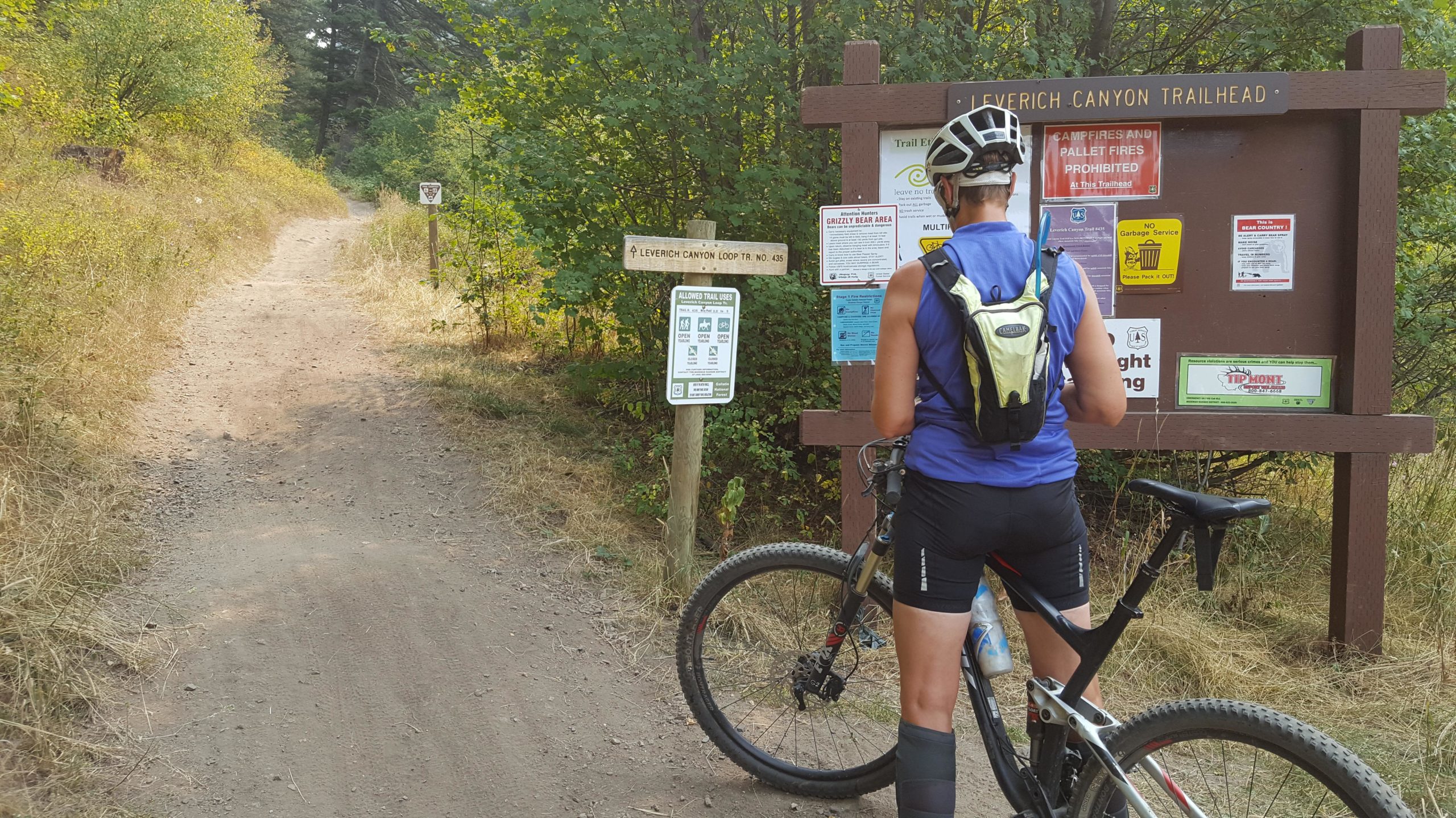 A person on a mountain bike stands near a trailhead sign for Leverich Canyon, surrounded by greenery and a dirt path. The sign displays various trail use regulations and safety information. The cyclist is wearing a blue tank top and a hydration pack and is checking the sign before starting their ride. Leverich Trail mountain bike trail.