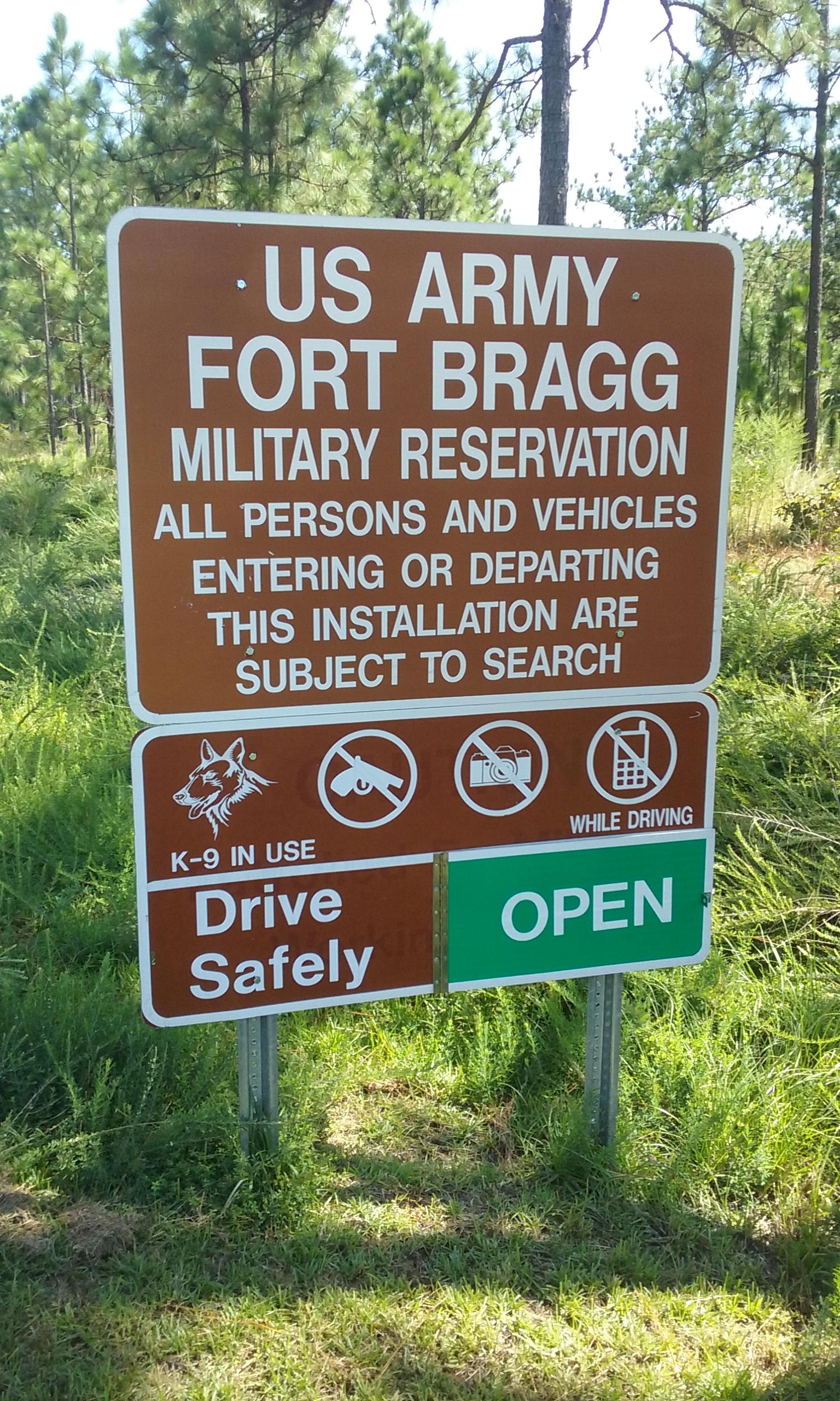 A brown and white sign at the entrance of Fort Bragg, US Army military reservation, indicating that all persons and vehicles entering or departing are subject to search. The sign features icons for K-9 use, and prohibits firearms, photography, and mobile phone use while driving. It also includes the phrase "Drive Safely" and states "OPEN." The background shows dense trees and grass. Smith Lake mountain bike trail.