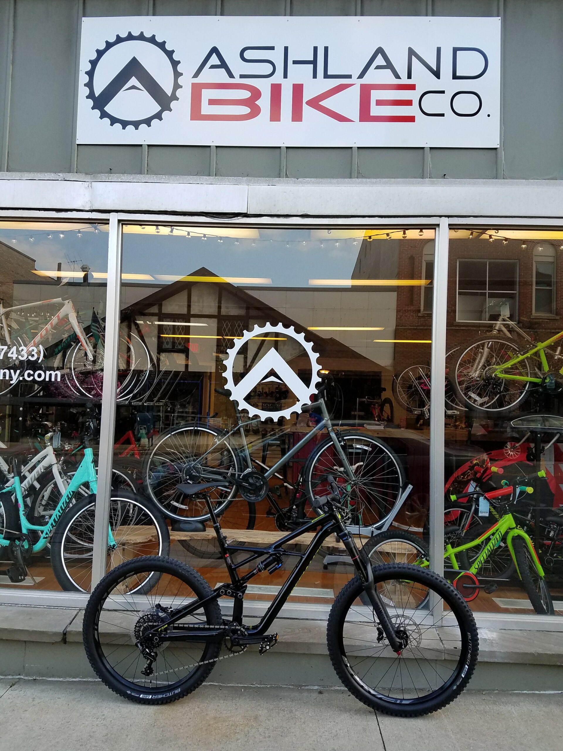 Alt text: A black mountain bike is positioned in front of the Ashland Bike Company storefront, which features a large sign above and a display of various bicycles in the window. The scene showcases the shop's bike offerings and signage.