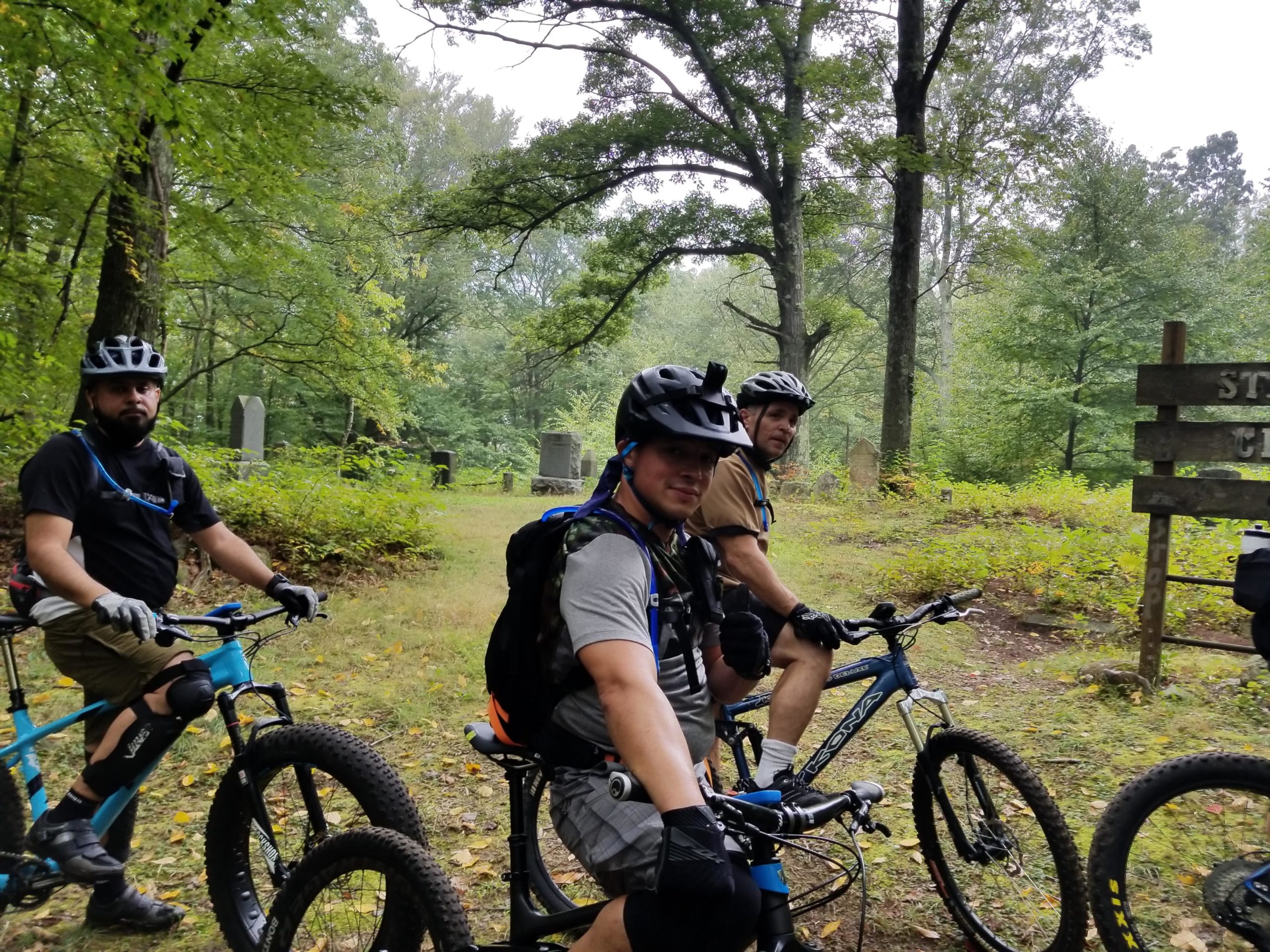 Three mountain bikers are posed with their bicycles in a wooded area near a trail sign. The environment features lush greenery and gravestones partially visible in the background. The men are wearing helmets and cycling gear, with two of them facing the camera and one giving a thumbs-up. The atmosphere is serene and slightly overcast. Wildcat Ridge NJ mountain bike trail.