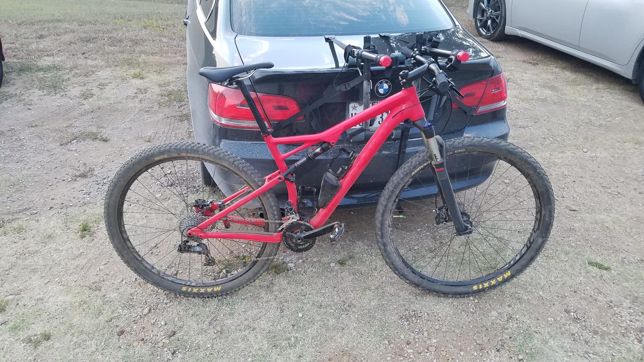 Specialized Epic Comp 29er: A red mountain bike is leaning against the rear of a black car in a gravel parking area. The bike's frame features a suspension system, and it has thick, knobby tires. A bike rack is mounted on the car, and the setting appears to be outdoors with grassy patches nearby.