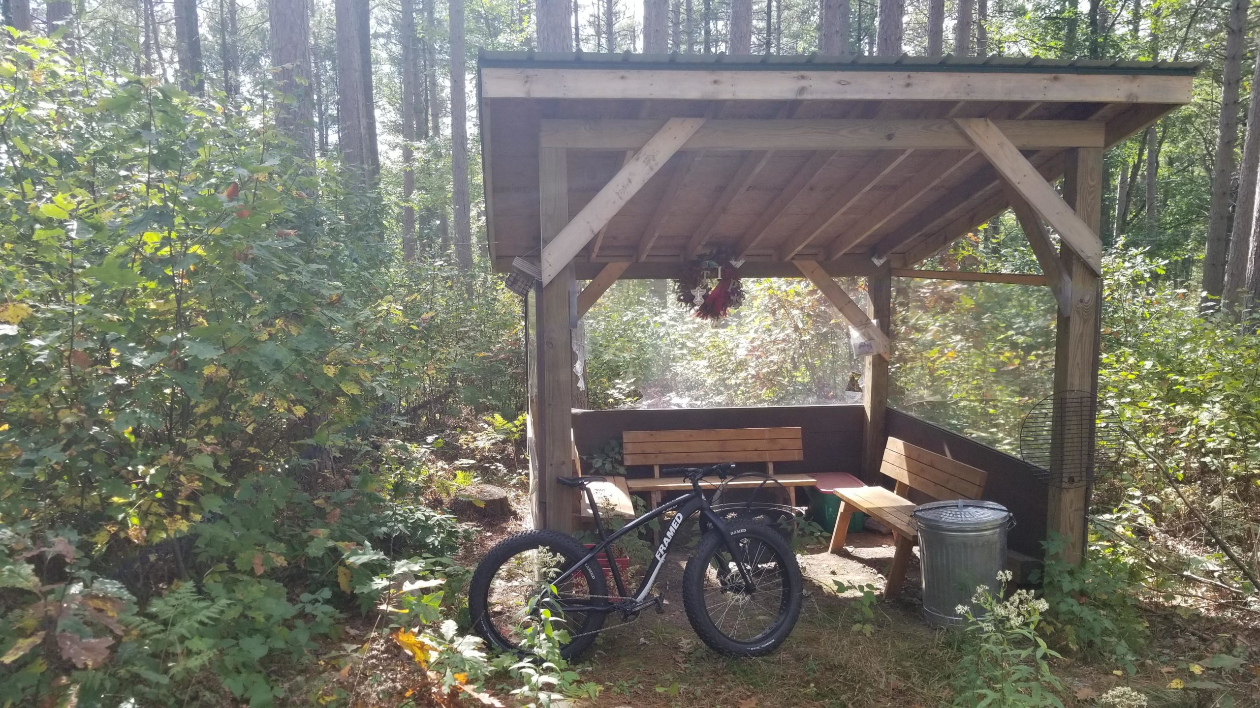 A wooden shelter with benches, set in a forested area surrounded by trees and greenery. A mountain bike is parked nearby, and there is a trash can next to the structure. Soft sunlight filters through the trees, creating a peaceful outdoor scene. Razorback Ridge mountain bike trail.