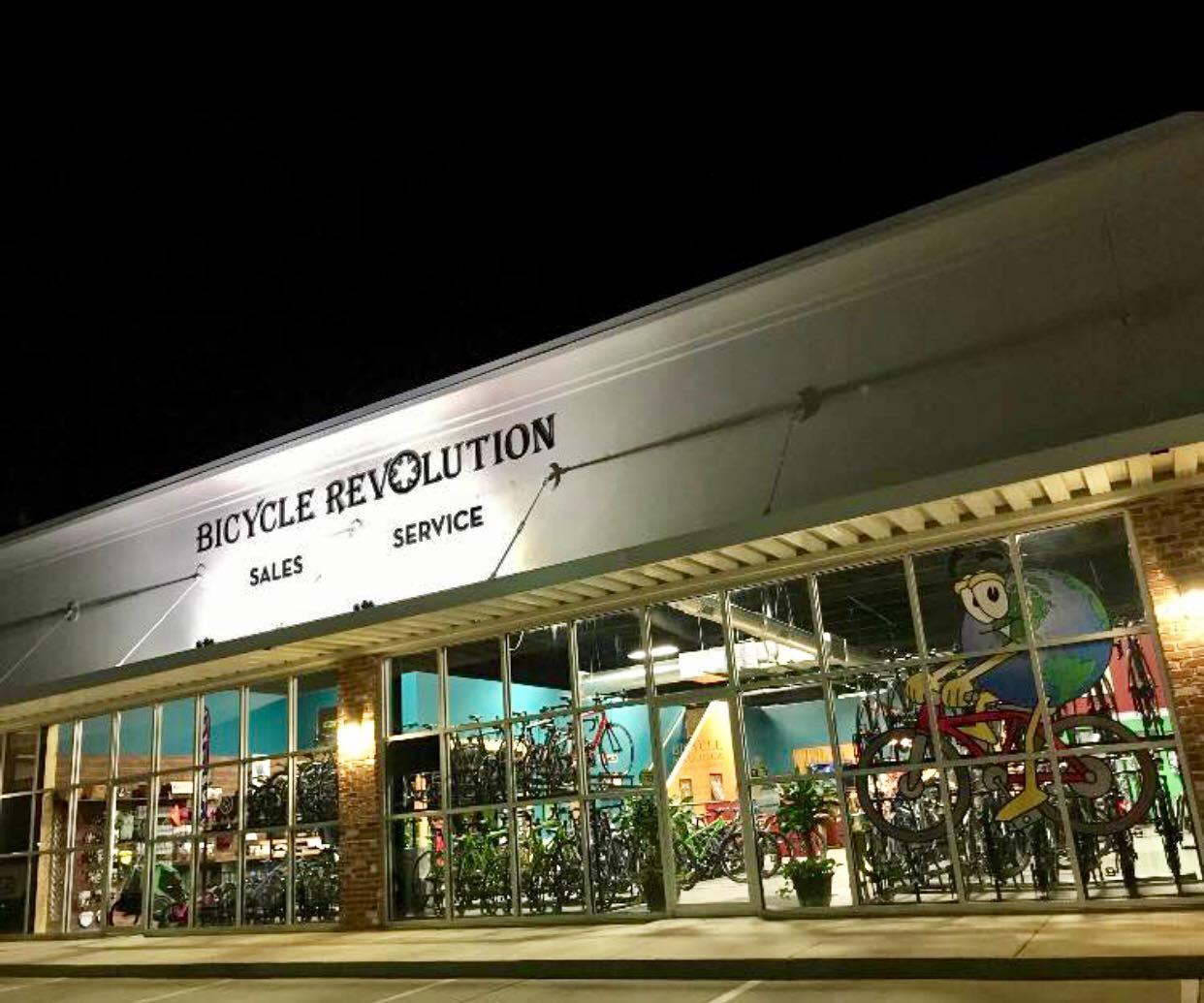 An exterior view of a bicycle shop, "Bicycle Revolution," at night. The building features large glass windows displaying various bicycles inside and a colorful mural of a cartoon character riding a bike. The signage prominently displays the words "Bicycle Revolution," "Sales," and "Service." The shop is illuminated from above, creating a welcoming atmosphere.