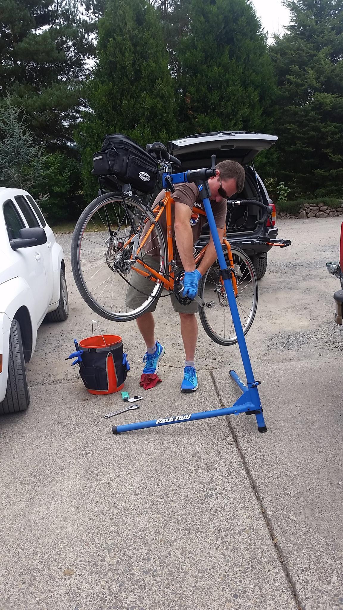 A person wearing gloves is working on a bicycle mounted on a blue repair stand in an outdoor setting. The scene includes a white car nearby, a black bag on the bike, and various tools in an orange bucket on the ground. Green trees are visible in the background.