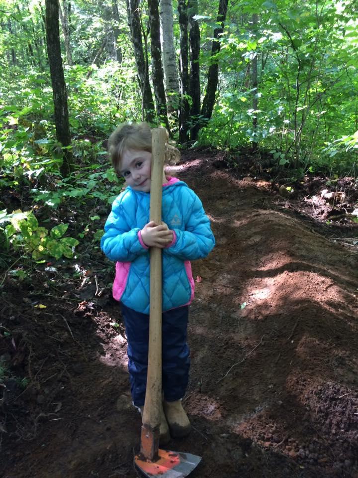A young girl in a blue jacket is standing in a wooded area, holding a shovel close to her body with a smile. She is surrounded by green foliage and dirt, indicating an outdoor activity or project. St-Côme mountain bike trail.