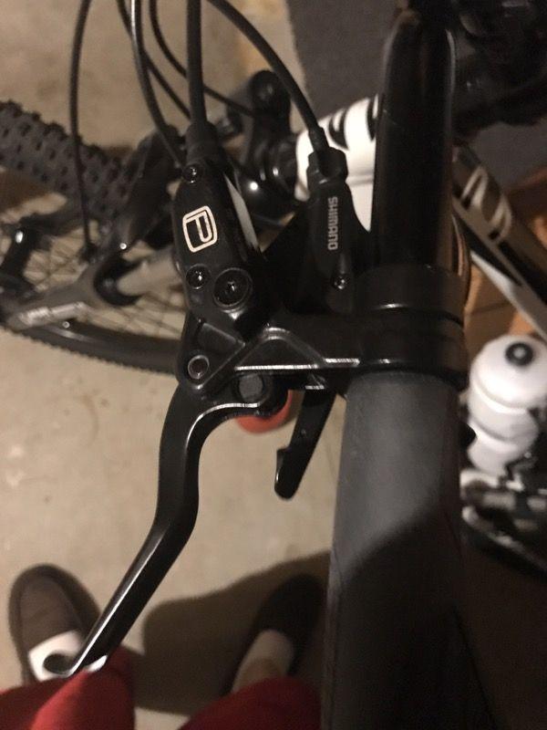 Cannondale Catalyst 2: Close-up image of a bicycle's brake lever mounted on the handlebars. The lever is black and features a recognizable Shimano logo. The background shows part of the bike frame and some footwear in the lower corner, suggesting the setting is indoors, possibly in a garage.