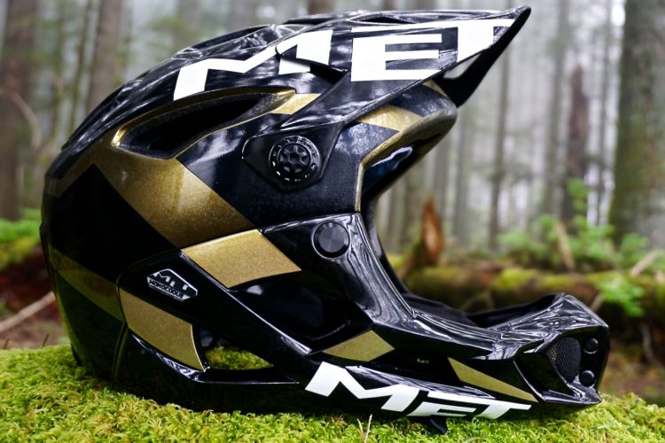 A black and gold mountain biking helmet resting on green moss, with a blurred forest background. The helmet features the brand name "MET" prominently displayed.
