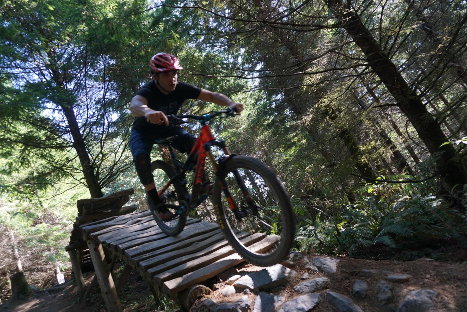 A mountain biker in a red helmet and a black shirt rides over a wooden plank bridge in a forested area, with trees and greenery visible in the background. The cyclist is in mid-air, demonstrating skill and agility on their bike. Galbraith Mountain mountain bike trail.