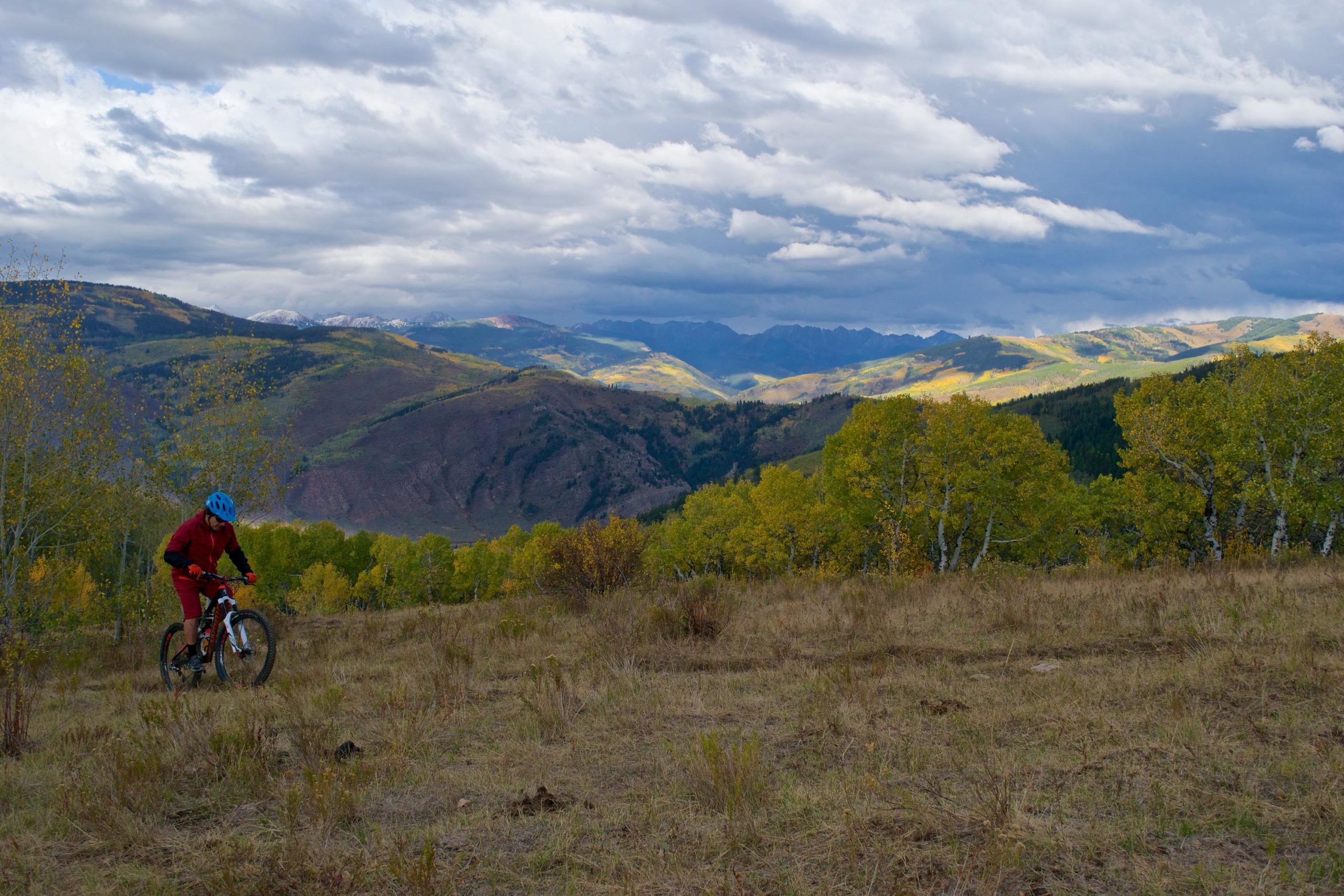 A person riding a mountain bike on a grassy hillside, surrounded by colorful autumn trees and majestic mountain scenery. The sky is partly cloudy, with patches of blue peeking through. Beaver Creek Ski Resort mountain bike trail.