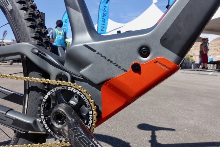 Close-up view of a high-performance mountain bike's drivetrain, featuring a carbon fiber crankset, gold chain, and an intricate suspension design. The bike's frame has a sleek gray finish with an orange accent, showcasing the brand name "Pardus." In the background, blurred figures and banners from an outdoor event can be seen.