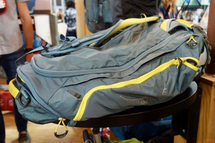 A blue and yellow outdoor backpack is displayed on a black circular table. The backpack features a zipper closure, side pockets, and adjustable straps. Blurred figures of people can be seen in the background, indicating a shopping or event environment.