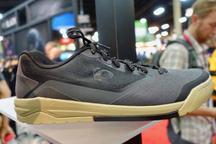 A close-up view of a grey athletic shoe displayed on a stand, featuring a sleek design with a black textured upper and a beige sole. The background includes blurred figures and displays, suggesting a busy trade show or exhibition environment.