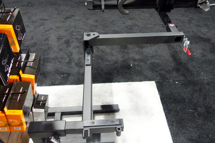 A black metal equipment stand with adjustable arms, positioned on a black carpeted floor. Nearby, several boxes of equipment are stacked, showcasing their orange and black packaging. The setup appears to be designed for display at a trade show or similar event.