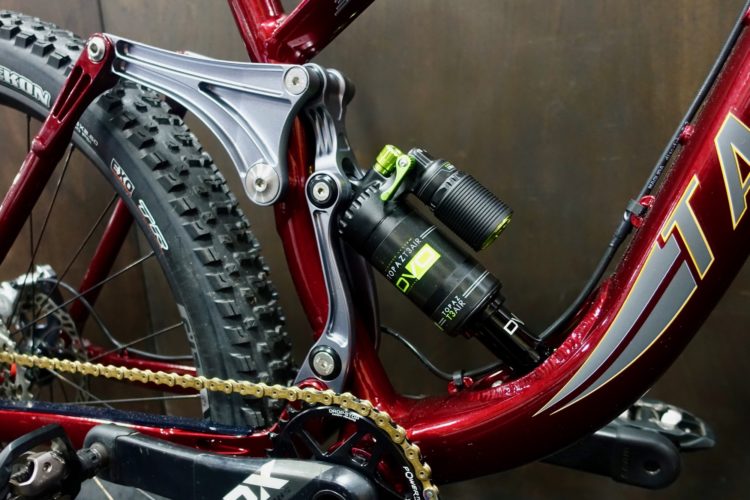 Close-up of a mountain bike's rear suspension system, featuring a dark red frame, a silver linkage, and a black shock absorber with green accents. The bike's chain and crankset are partially visible, showcasing its design for off-road performance. The background is a dark, textured surface.