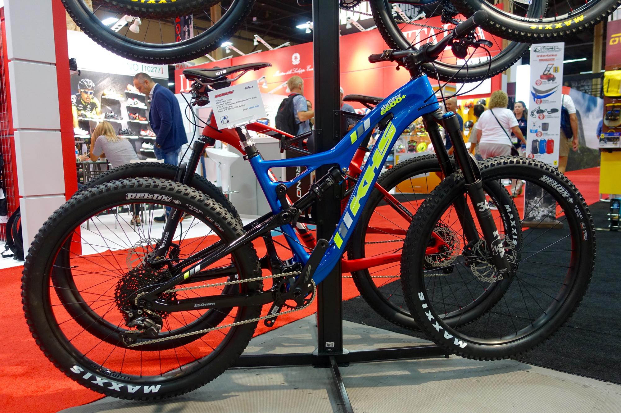 KHS sixfifty 6600: A display of two mountain bikes positioned on a rack, one in blue and the other in red. The bikes feature thick tires and modern design elements. In the background, there are blurred figures of people interacting, with various colorful merchandise displayed at an indoor exhibit or trade show. The setting has a red carpet and contrasting dark floor.