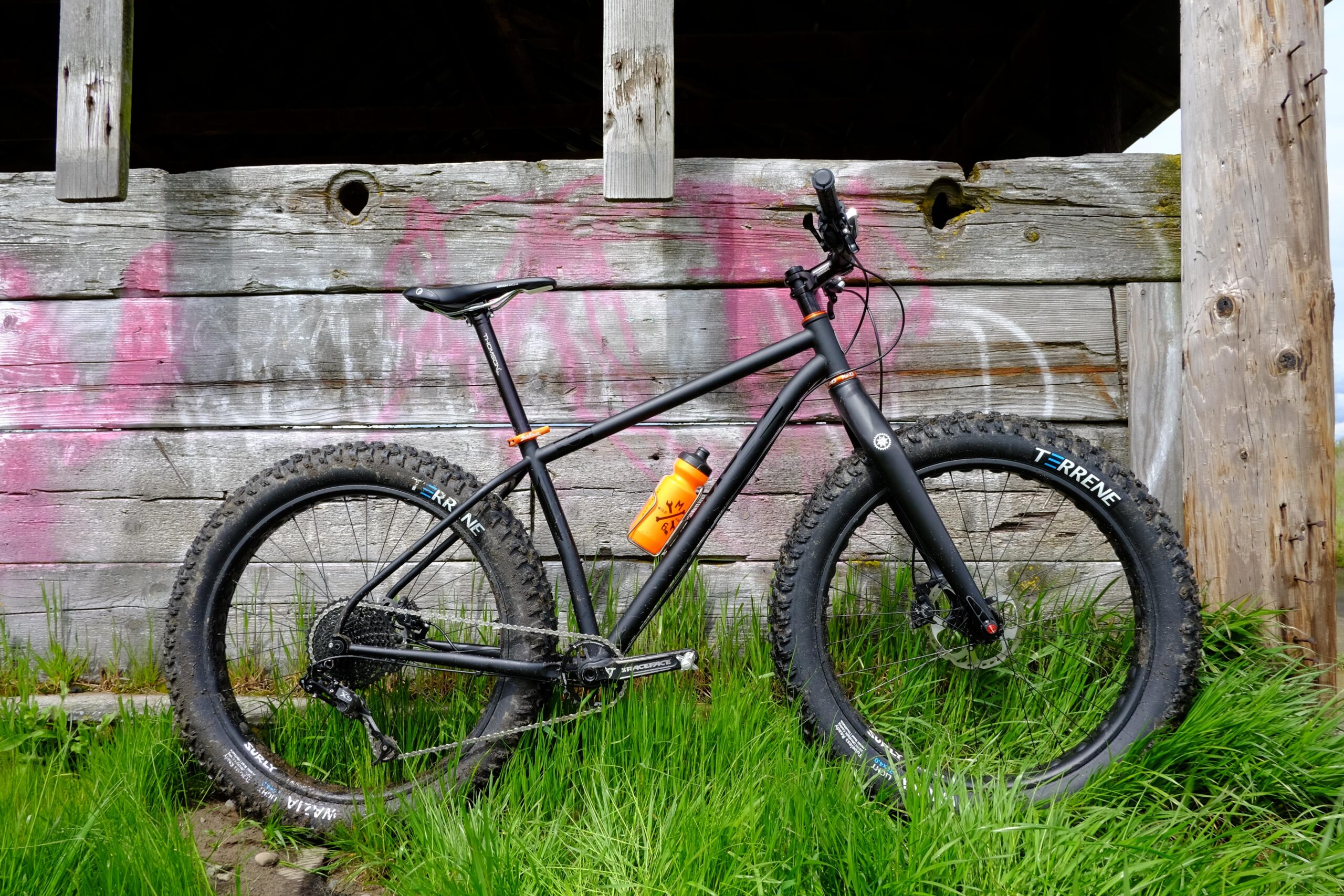 A black mountain bike with wide tires is leaning against a weathered wooden structure, partially covered in pink graffiti. An orange water bottle is attached to the bike frame, and lush green grass surrounds the base of the bike.
