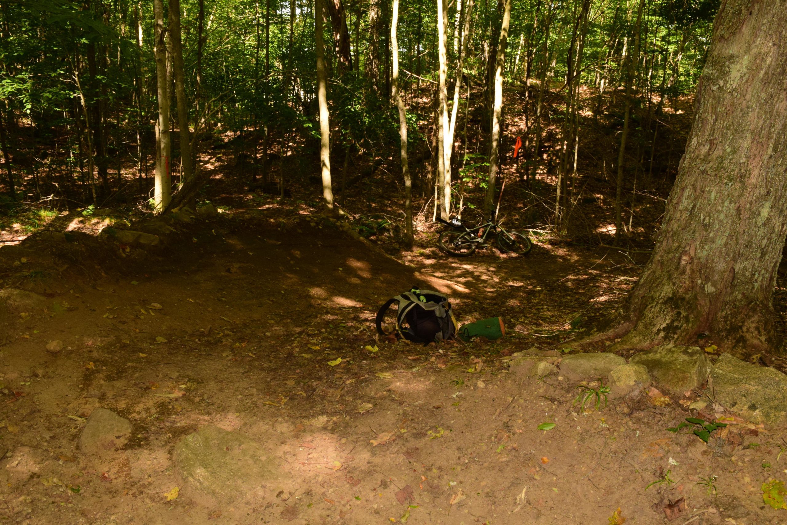 A wooded trail with a dirt path leading downward, surrounded by tall trees and leafy undergrowth. A black backpack is visible on the ground, and in the background, a bicycle is resting against a tree. Sunlight filters through the canopy, creating dappled light on the trail. Mount Pisgah II: the left side mountain bike trail.