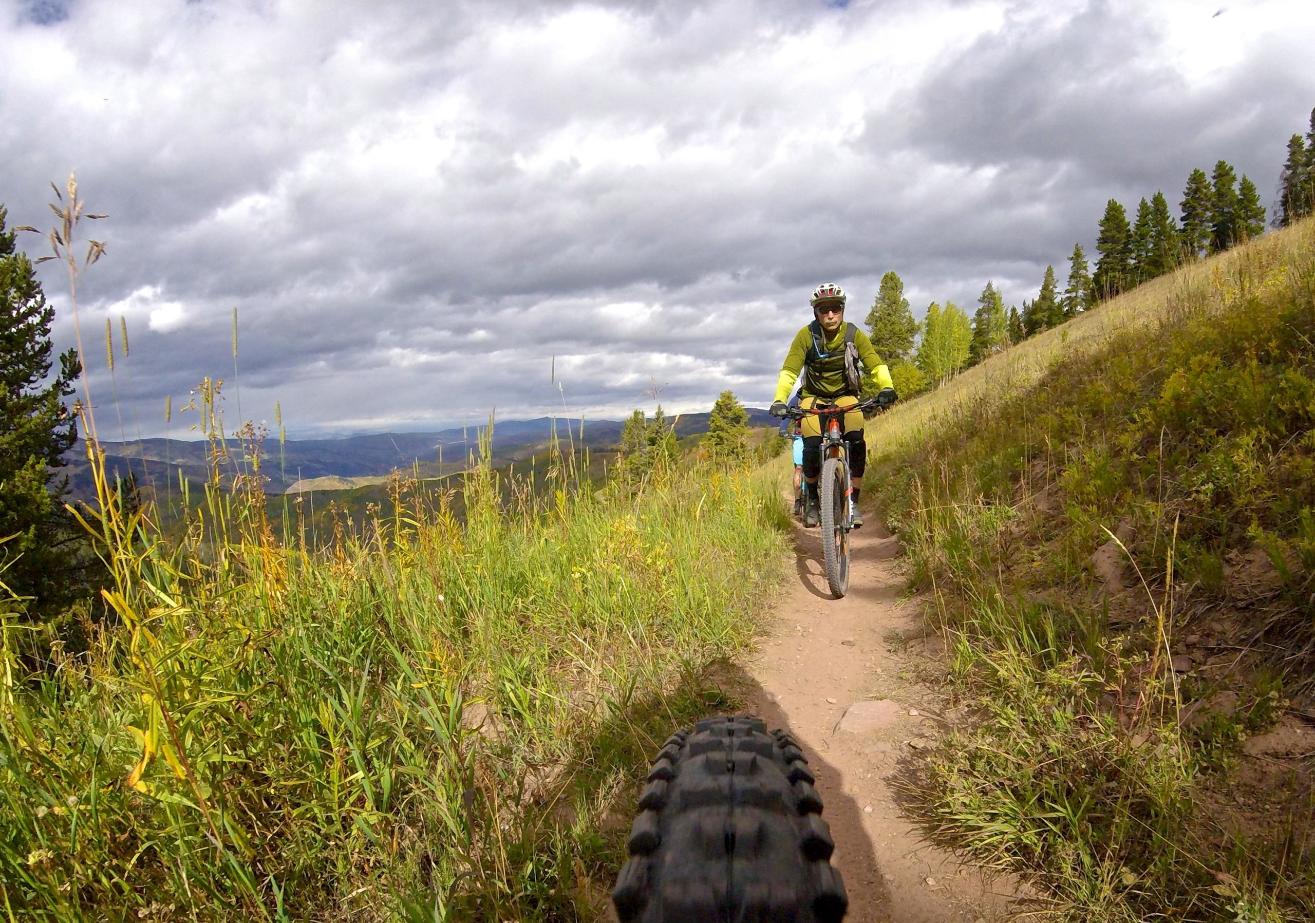 Mountain biker riding on a dirt trail surrounded by tall grass and wildflowers, with a scenic view of hills and cloudy sky in the background. Vail Mountain Bike Park mountain bike trail.