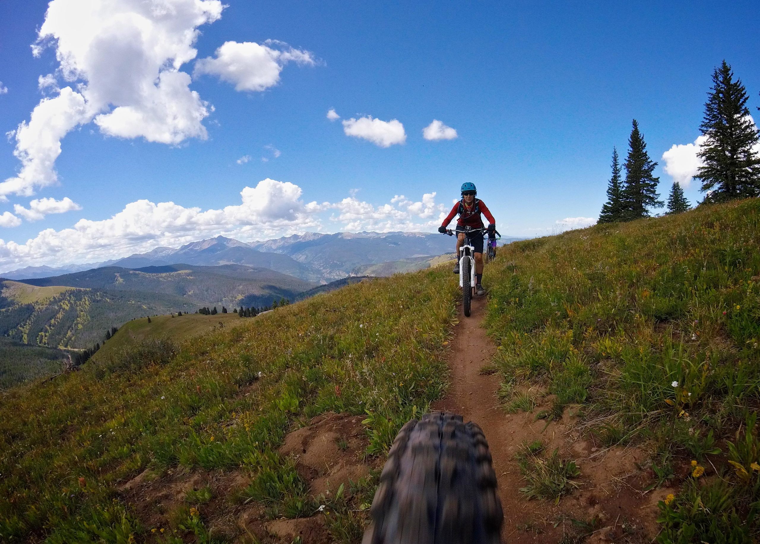 A mountain biker riding along a singletrack trail with scenic mountain views in the background, featuring green grass and wildflowers in the foreground. The sky is bright blue with fluffy white clouds, and a second biker is visible ahead on the path. Two Elk via Vail Pass mountain bike trail.