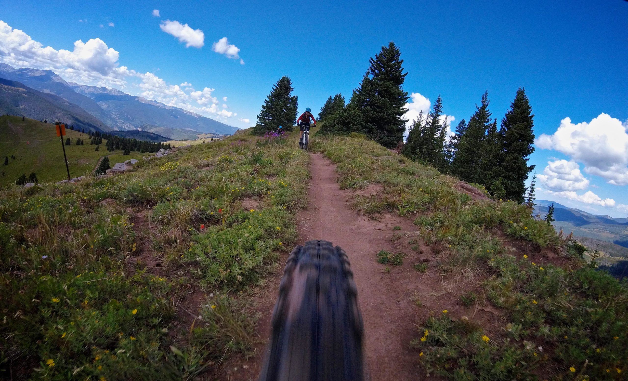 A mountain biking trail winding through green meadows and pine trees, with a rider visible in the distance against a backdrop of mountains and blue sky with fluffy clouds. The view includes a close-up of a bike tire in motion along the dirt path. Two Elk via Vail Pass mountain bike trail.