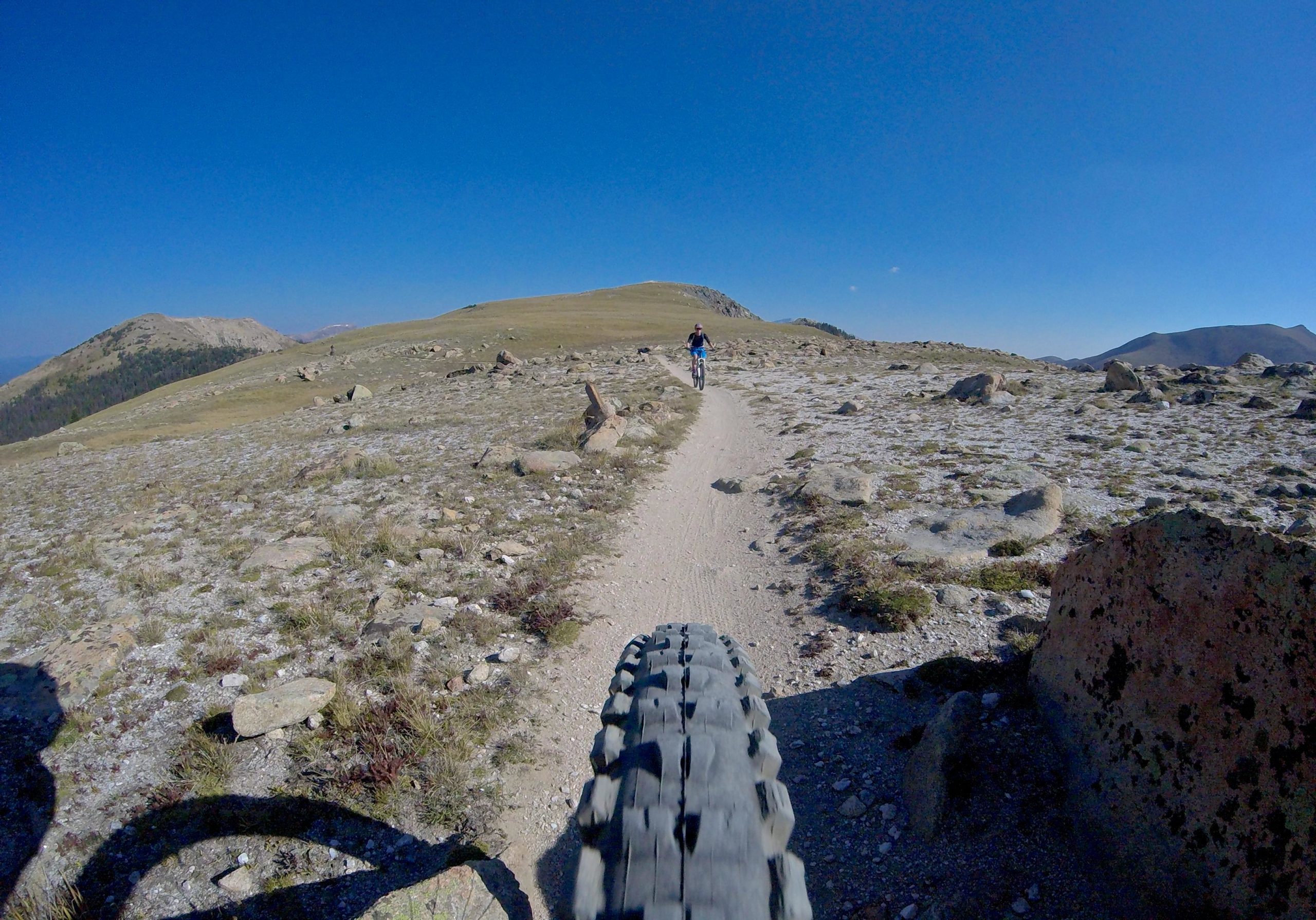 A mountain biker rides along a gravel path through a rocky landscape, surrounded by hills under a clear blue sky. The foreground features a close-up view of the bike tire on the trail, while the distant rider is seen pedaling ahead on the winding track. Monarch Crest Trail mountain bike trail.