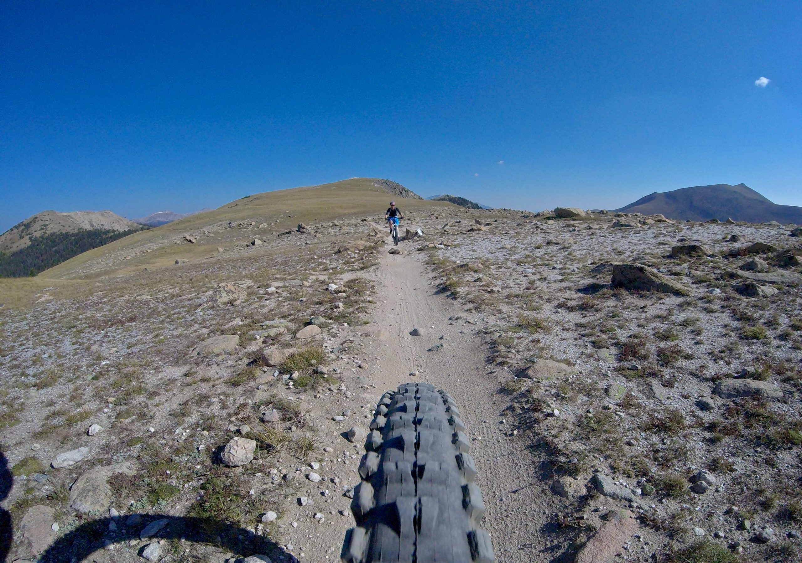 A view from the perspective of a mountain bike tire on a narrow dirt trail, with a cyclist visible ahead on a rocky terrain under a clear blue sky. Rolling hills and distant mountains are visible in the background. Monarch Crest Trail mountain bike trail.