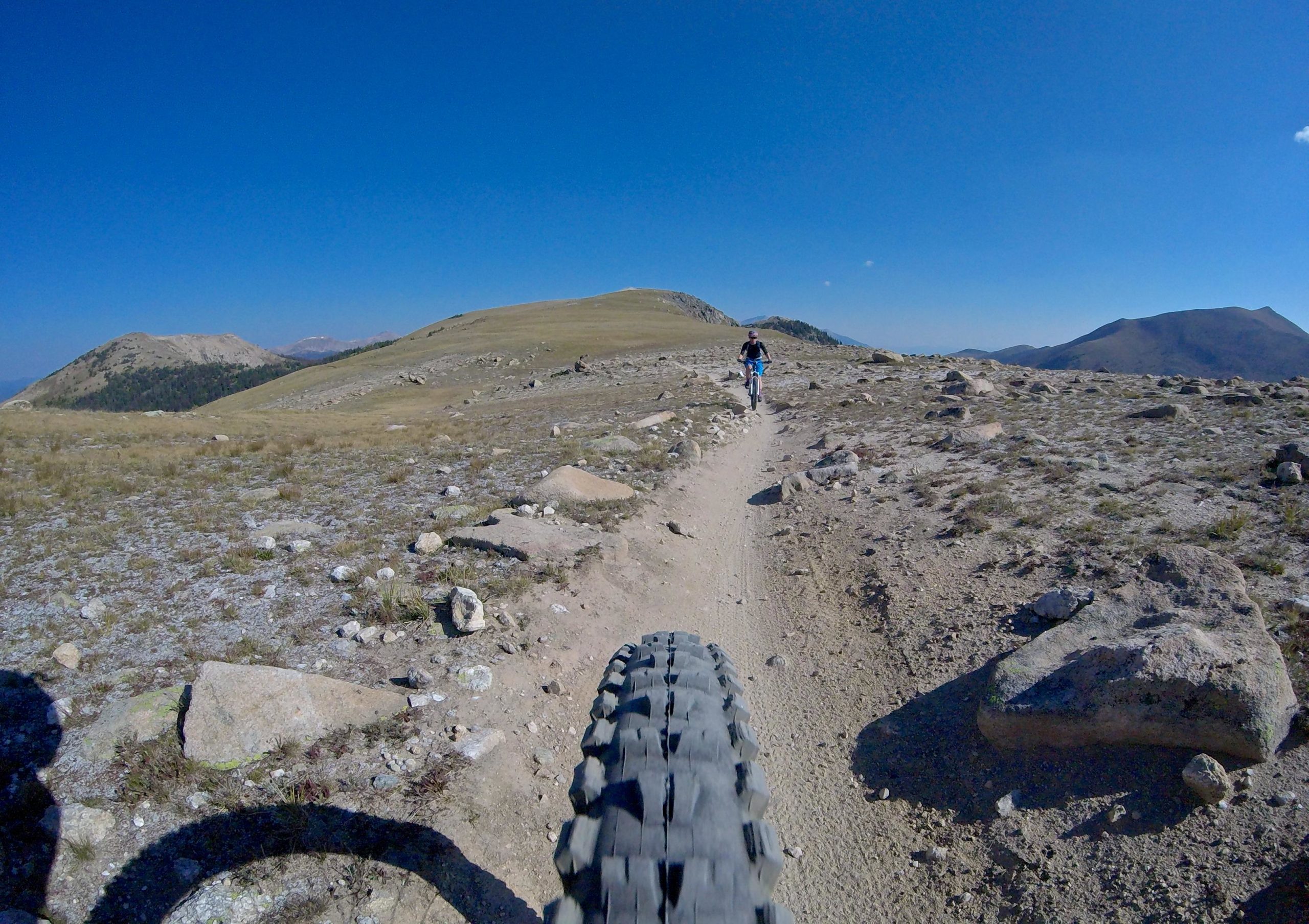 A mountain biker riding on a dirt trail in a mountainous landscape, with rocky terrain and blue skies in the background. The image is taken from a low angle, focusing on the bike's front tire and the path ahead. Monarch Crest Trail mountain bike trail.