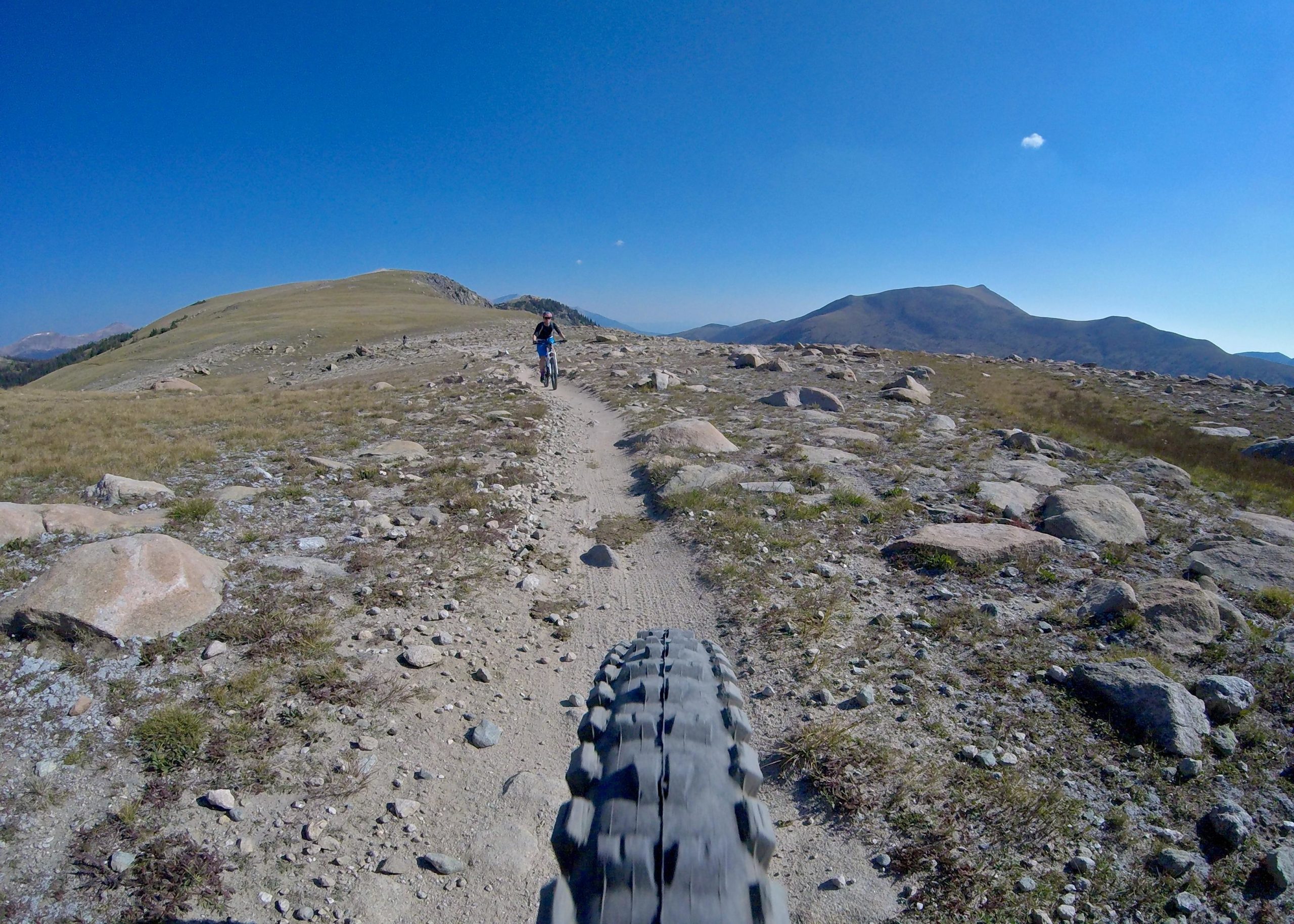 A mountain biker riding on a rocky trail in a mountainous landscape under a clear blue sky. The perspective is from the front of the bike, showing the tire in the foreground and the rider in the distance. Monarch Crest Trail mountain bike trail.