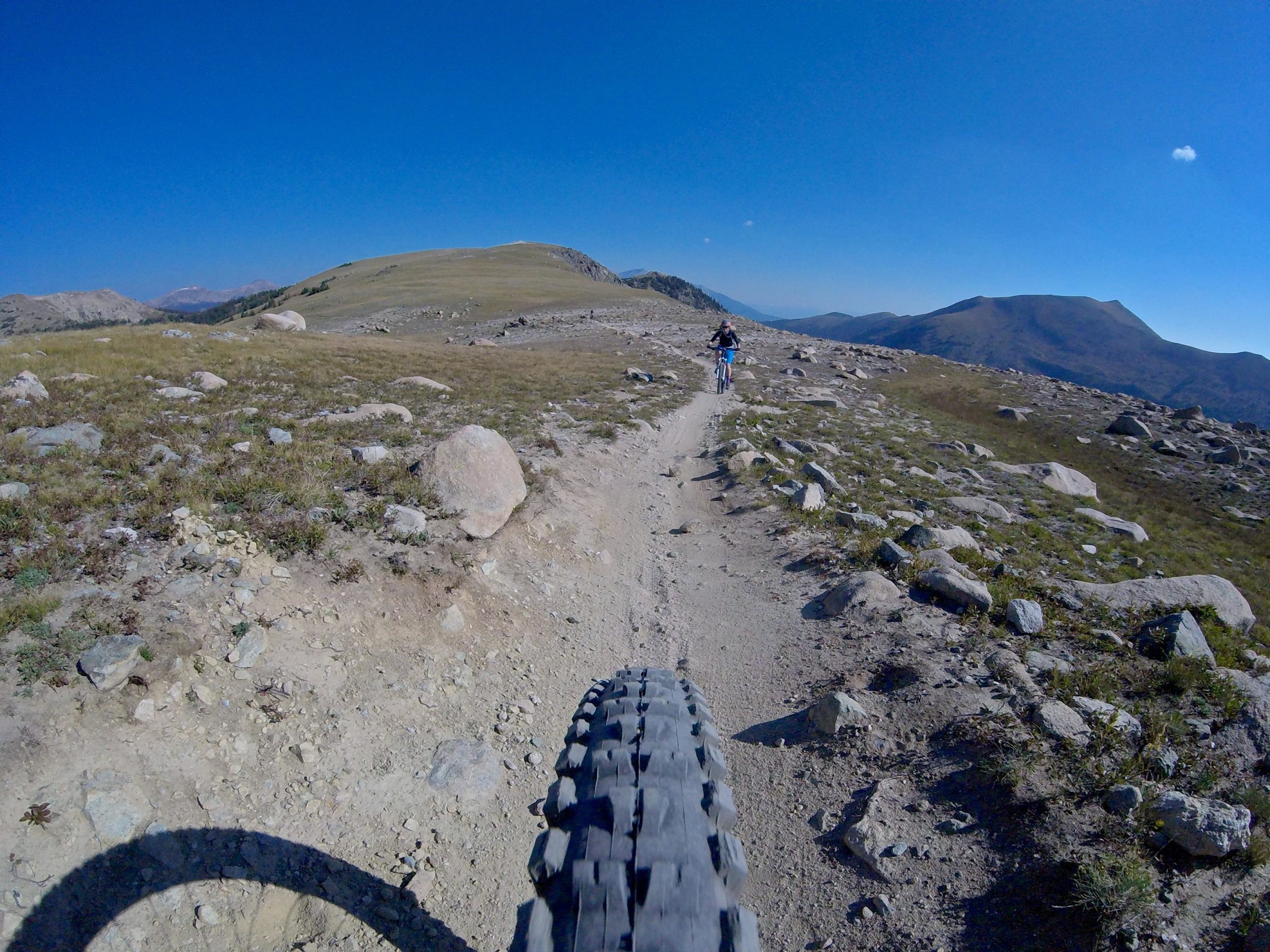 A mountain biker riding along a rocky and uneven trail in a vast landscape with blue sky and distant mountains. The view shows the bike tire in the foreground, highlighting the rough terrain and natural setting. Monarch Crest Trail mountain bike trail.