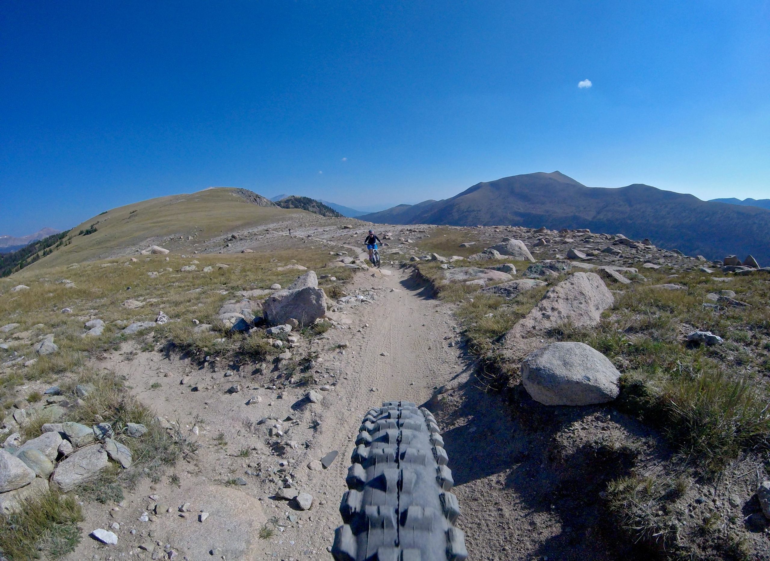 A mountain biking trail with a view of rolling hills and rocky terrain under a clear blue sky. The image shows the perspective from a bike, focusing on the tire and path ahead, with another cyclist in the distance. Monarch Crest Trail mountain bike trail.