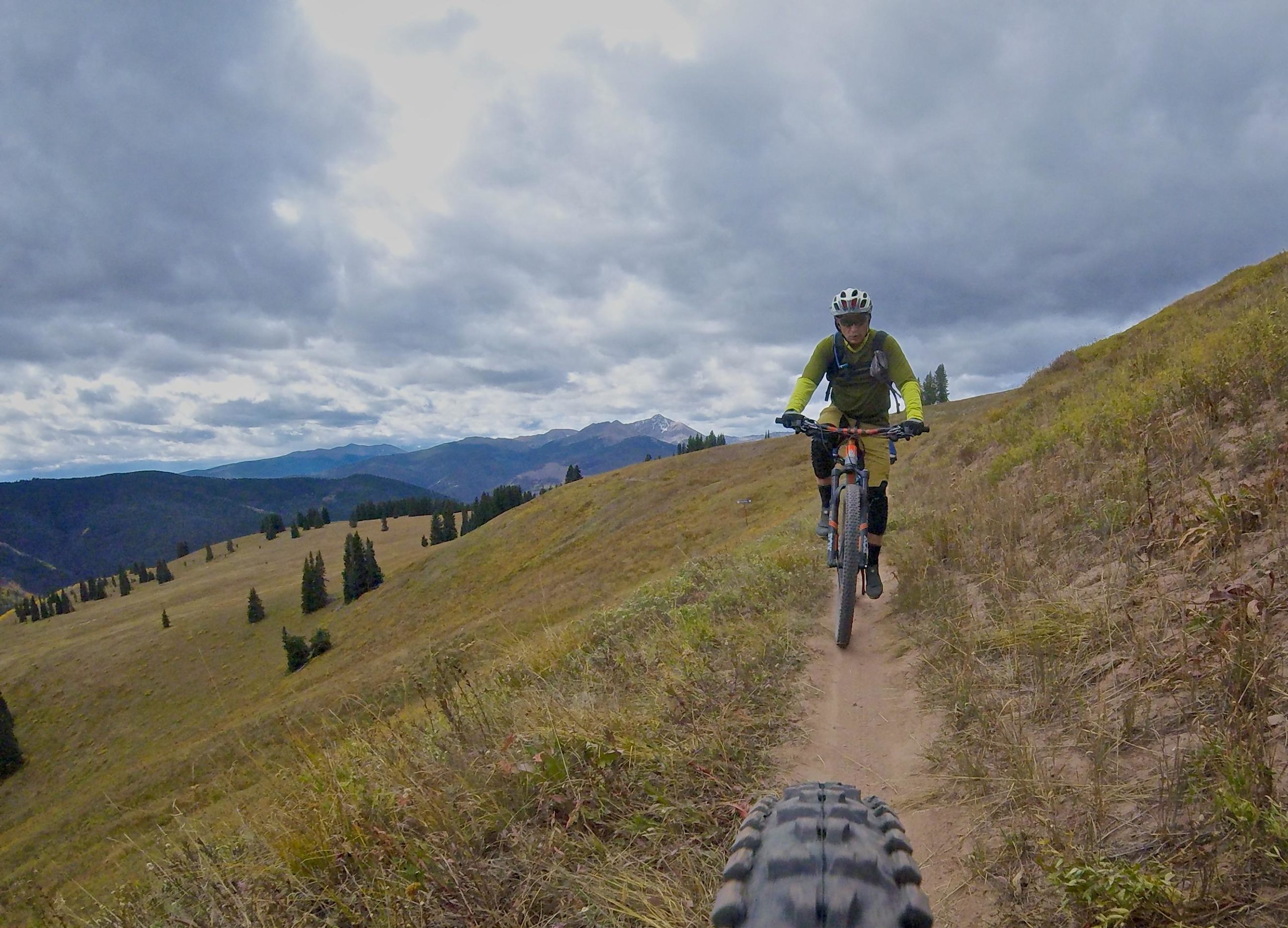 A mountain biker in a green long-sleeve shirt and helmet rides along a narrow dirt path on a hillside, surrounded by grassy terrain and pine trees. The background features mountains under a cloudy sky, creating a scenic outdoor atmosphere. Vail Mountain Bike Park mountain bike trail.