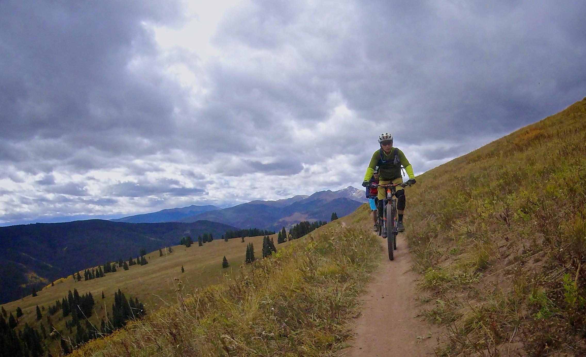 A cyclist riding along a narrow dirt trail in a mountainous landscape, with grassy fields and pine trees in the foreground. The backdrop features rolling hills and distant mountains under a cloudy sky. Vail Mountain Bike Park mountain bike trail.