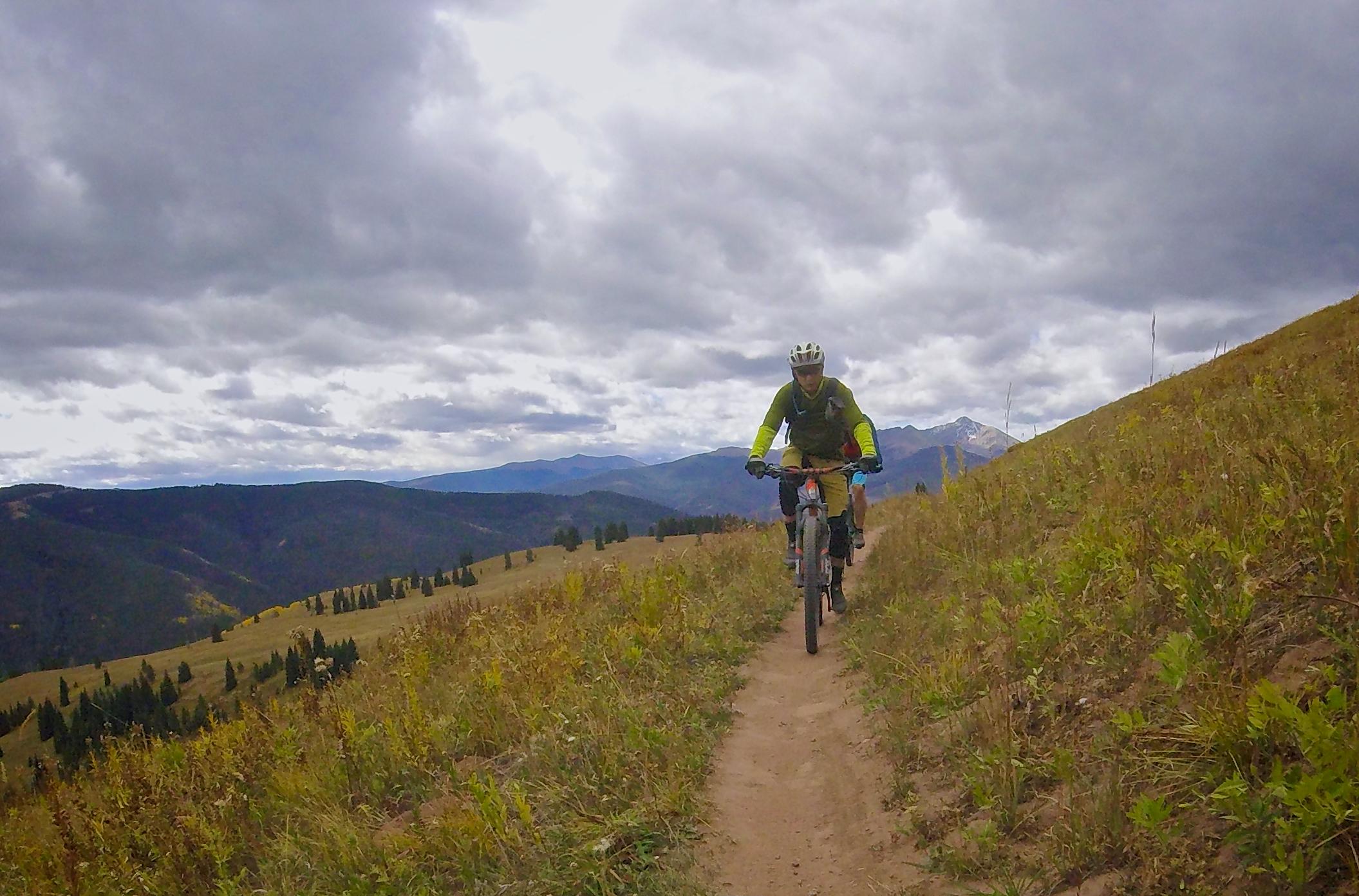 A cyclist riding a mountain bike along a dirt trail through grassy terrain, with mountains and cloudy skies in the background. Vail Mountain Bike Park mountain bike trail.