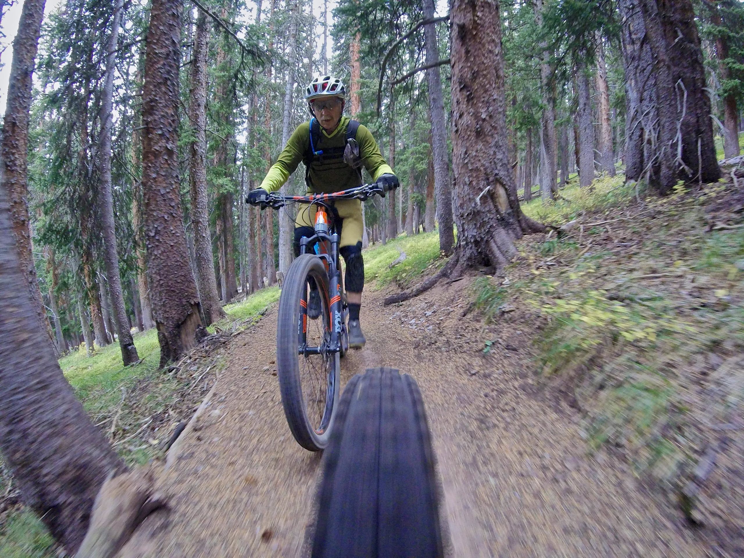 Mountain biker riding on a dirt trail surrounded by tall trees in a forest. The cyclist is wearing a helmet and protective gear, focused and maneuvering through the path, with the front tire of the bike visible in the foreground. The scene captures the essence of outdoor adventure and the beauty of nature. Vail Mountain Bike Park mountain bike trail.