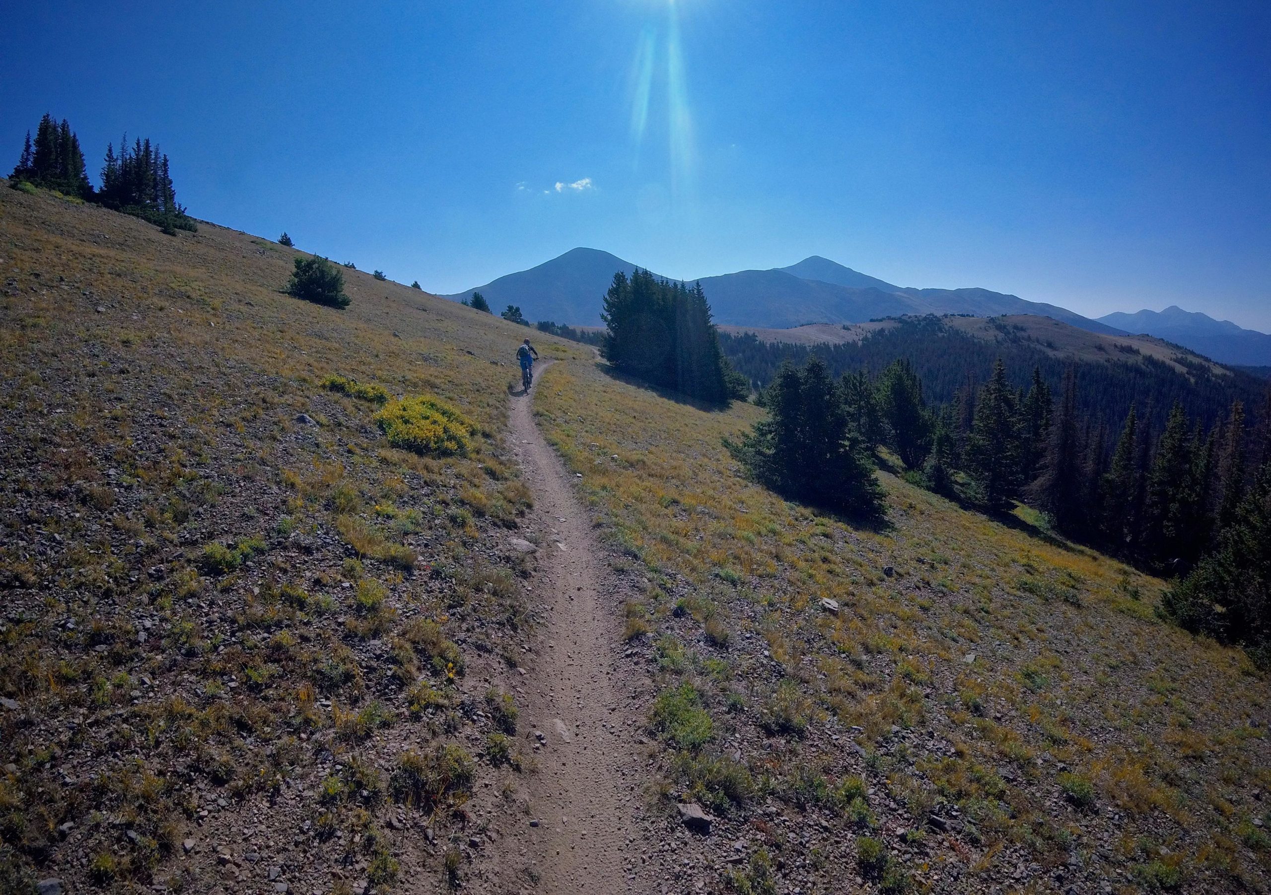 A lone hiker walking along a dirt trail on a grassy hillside, with mountains in the background under a clear blue sky. The landscape features patches of greenery and rocky terrain illuminated by sunlight. Monarch Crest Trail mountain bike trail.