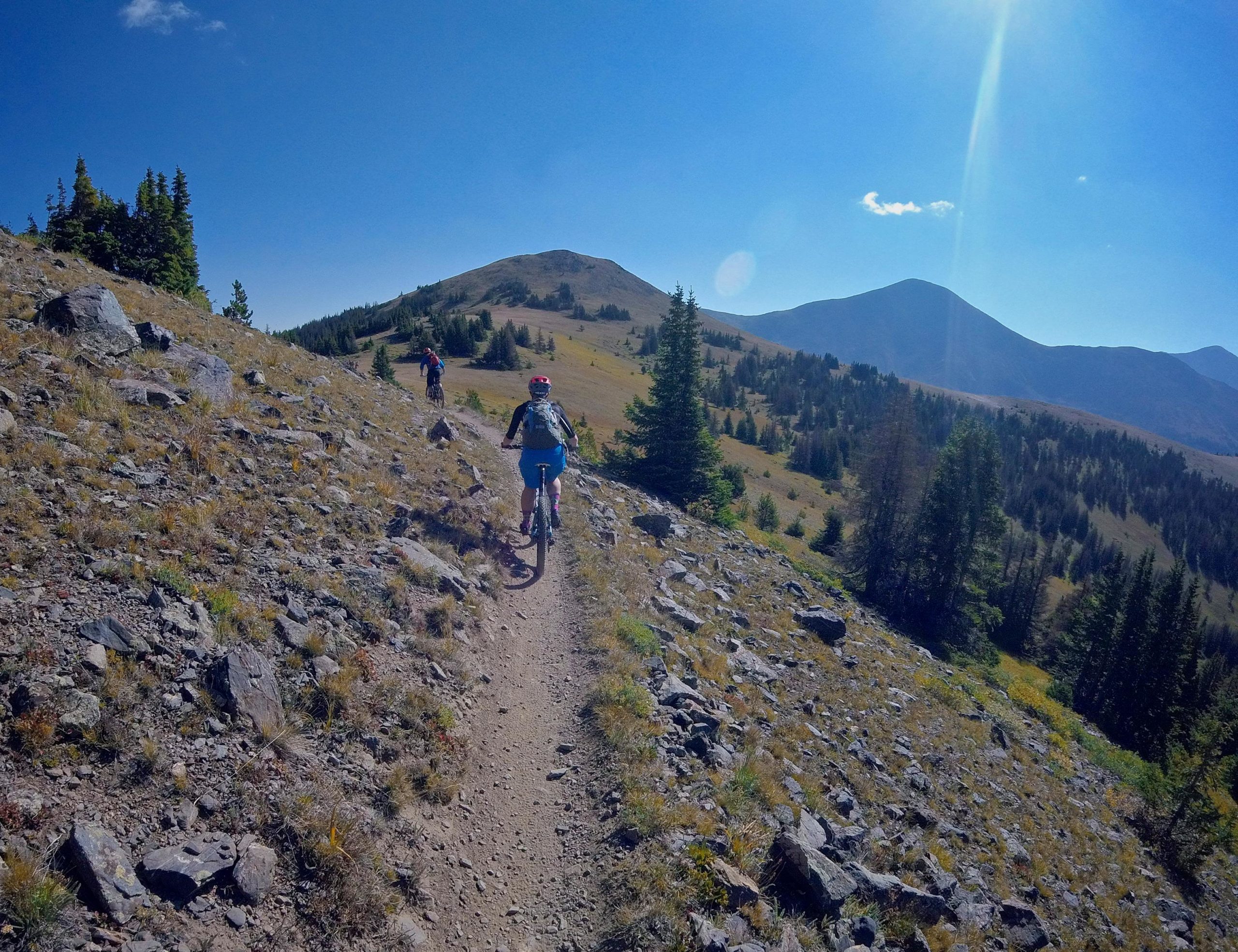 Mountain bikers riding along a rocky trail surrounded by trees and mountains under a bright blue sky. Monarch Crest Trail mountain bike trail.