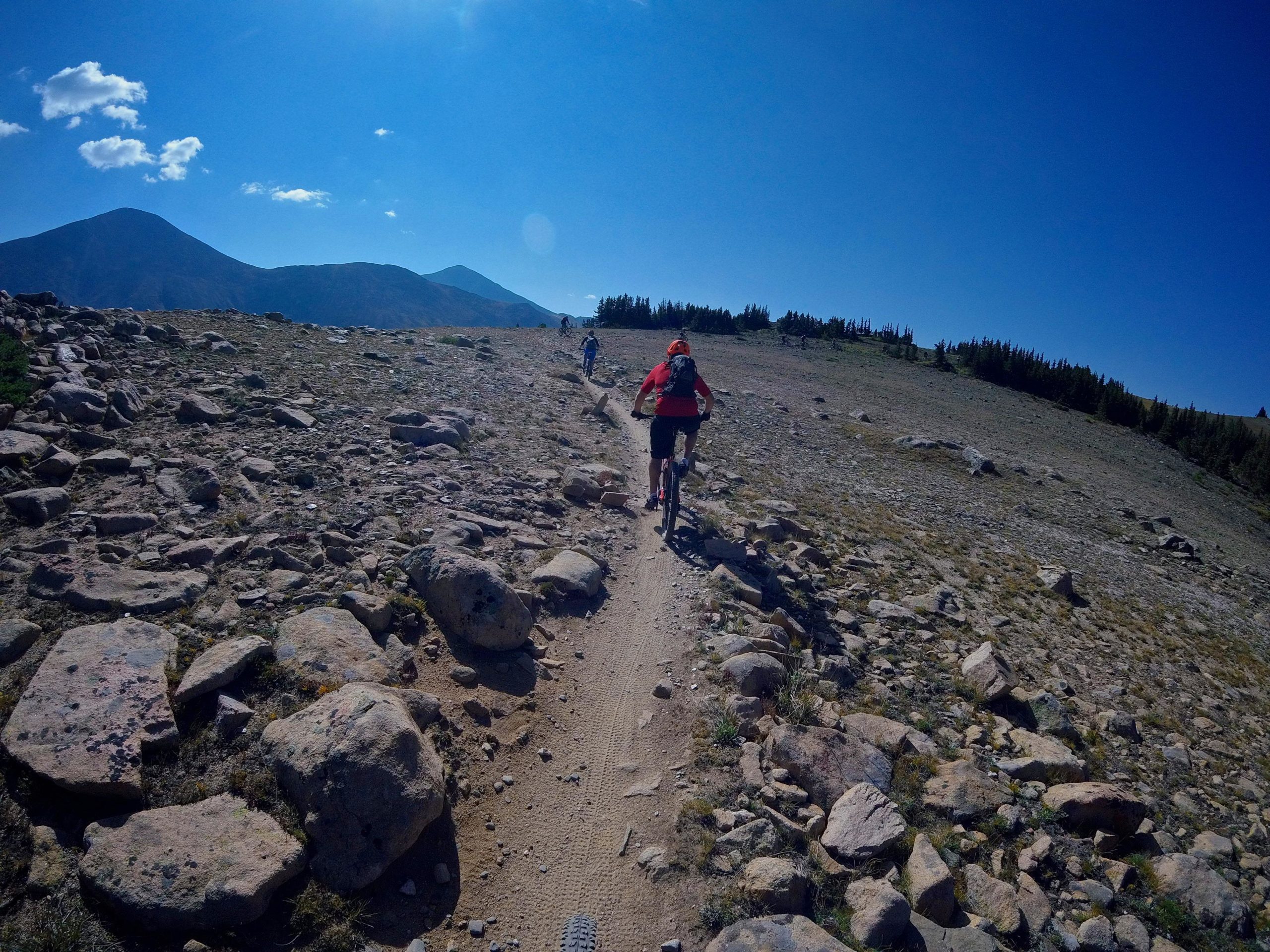 A mountain biker rides along a rocky trail in a mountainous landscape under a clear blue sky. The path is surrounded by boulders and sparse vegetation, with distant mountains visible in the background. Monarch Crest Trail mountain bike trail.