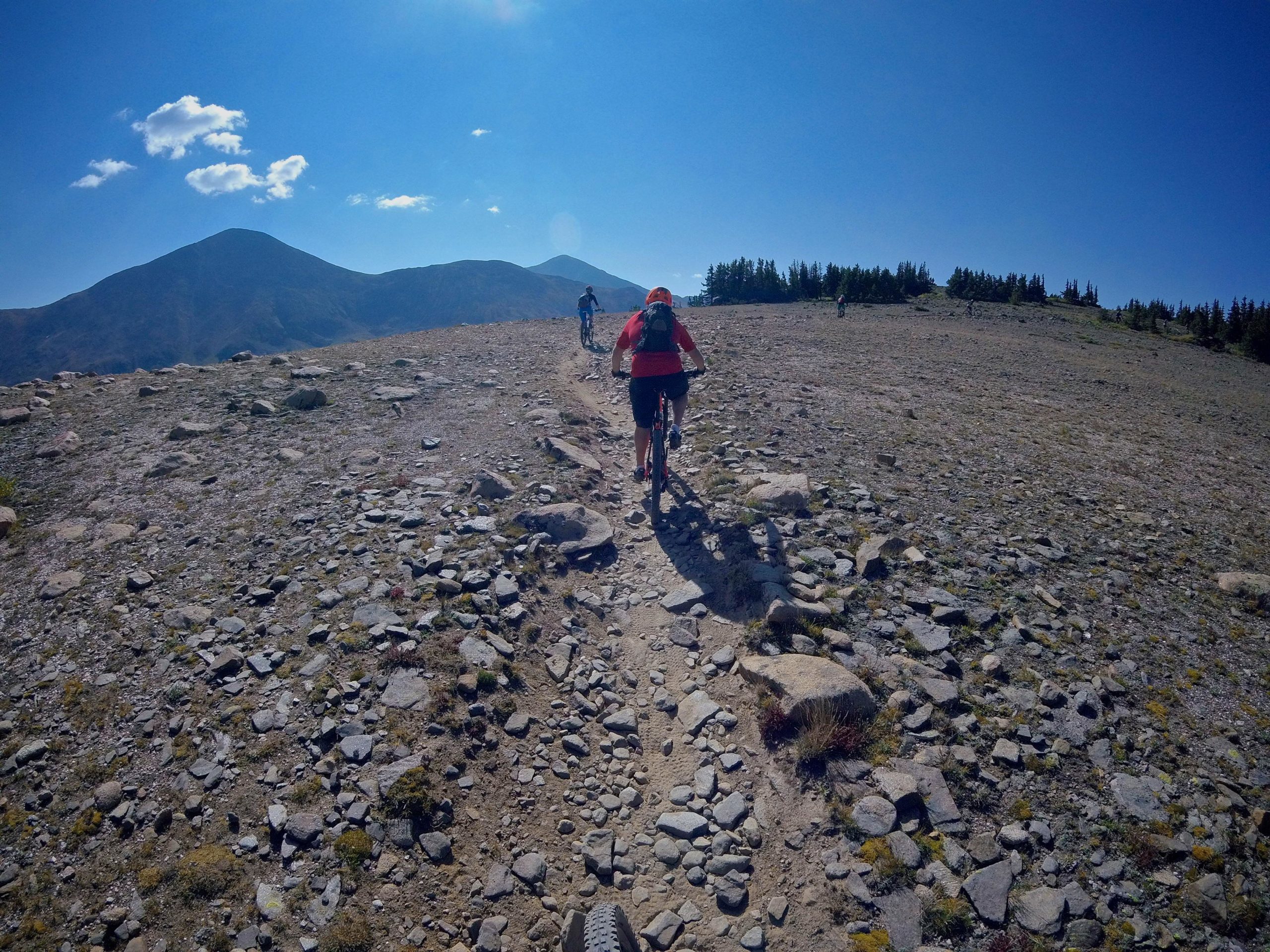 Mountain bikers ride on a rocky trail with mountains in the background under a clear blue sky. The scene captures a remote and rugged landscape, illustrating the adventure of outdoor cycling. Monarch Crest Trail mountain bike trail.