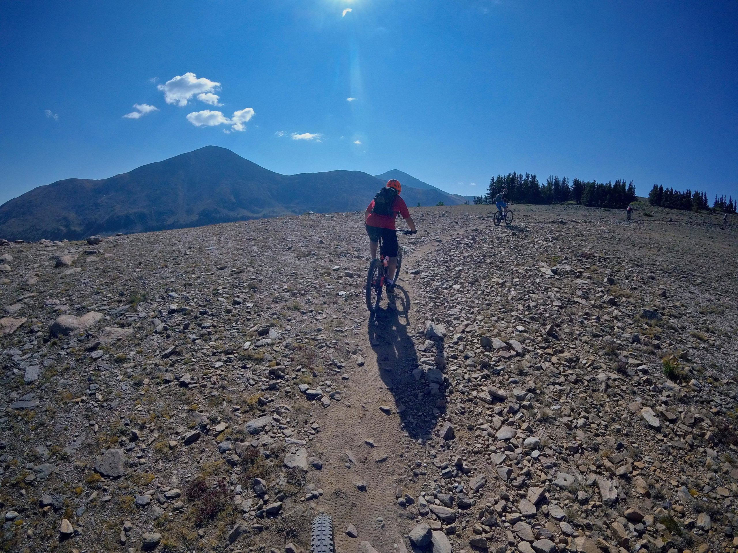 A group of mountain bikers riding on a rocky trail in a mountainous landscape under a bright blue sky. The foreground features loose stones and a dirt path, while distant mountains rise against the backdrop of the sky with a few clouds. Monarch Crest Trail mountain bike trail.