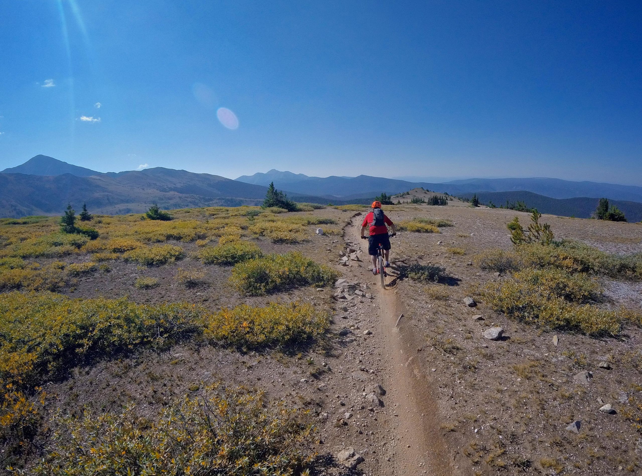 A cyclist riding along a dirt trail through a mountainous landscape, surrounded by low-lying vegetation and rocky terrain, under a clear blue sky. The background features rolling hills and distant mountain peaks. Monarch Crest Trail mountain bike trail.