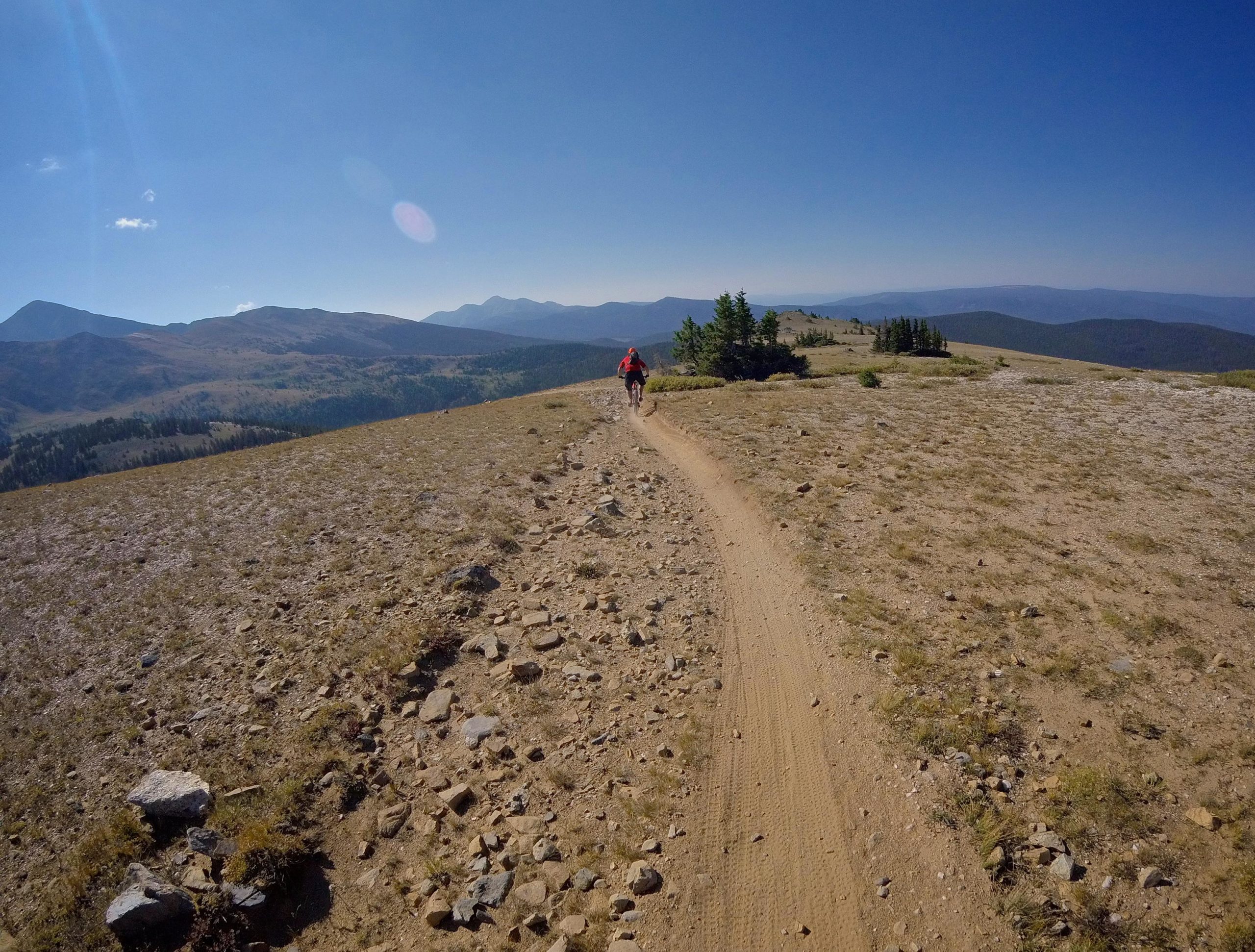 A person wearing a red jacket biking along a narrow, rocky trail on a mountain ridge, surrounded by expansive views of rolling hills and distant mountains under a clear blue sky. Monarch Crest Trail mountain bike trail.