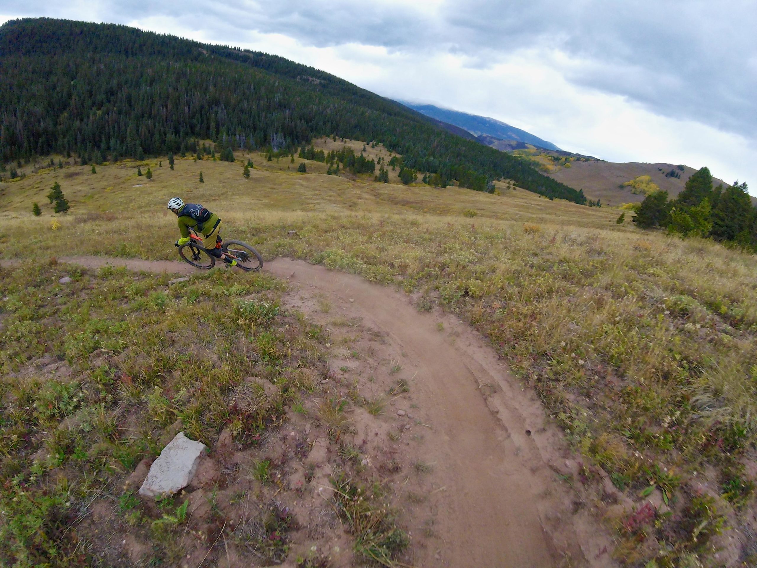 A mountain biker navigating a winding dirt trail surrounded by grassy terrain and forested hills under a cloudy sky. Two Elk via Vail Pass mountain bike trail.