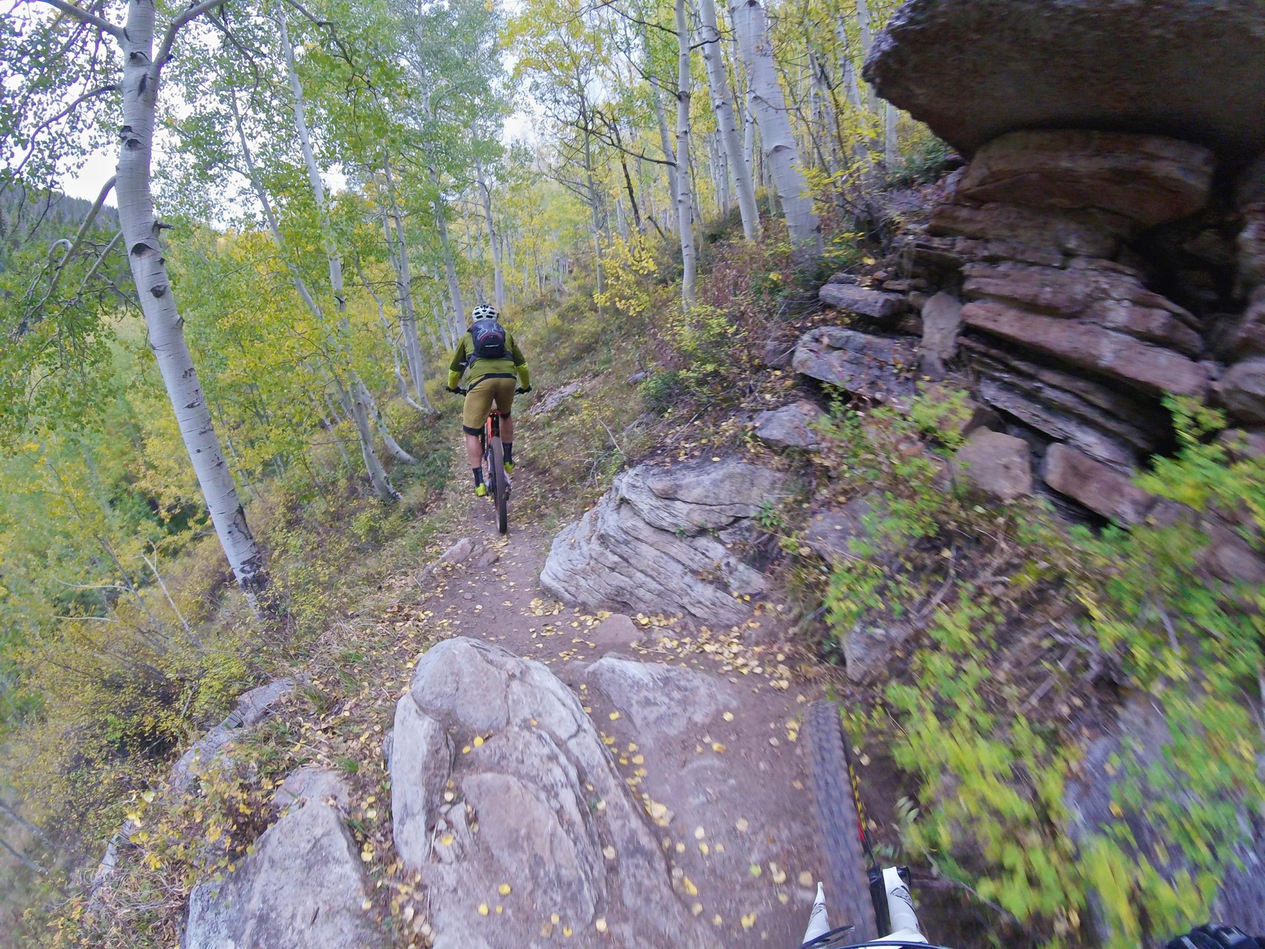 A mountain biker navigates a rocky trail surrounded by green and yellow trees, set against a backdrop of rocky terrain and an overhanging boulder. Two Elk via Vail Pass mountain bike trail.